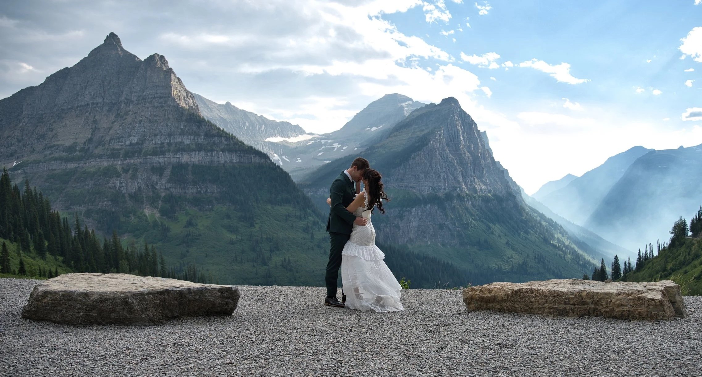 A bride and groom embrace in front of mountain scenery with evergreen trees and a cloudy sky.