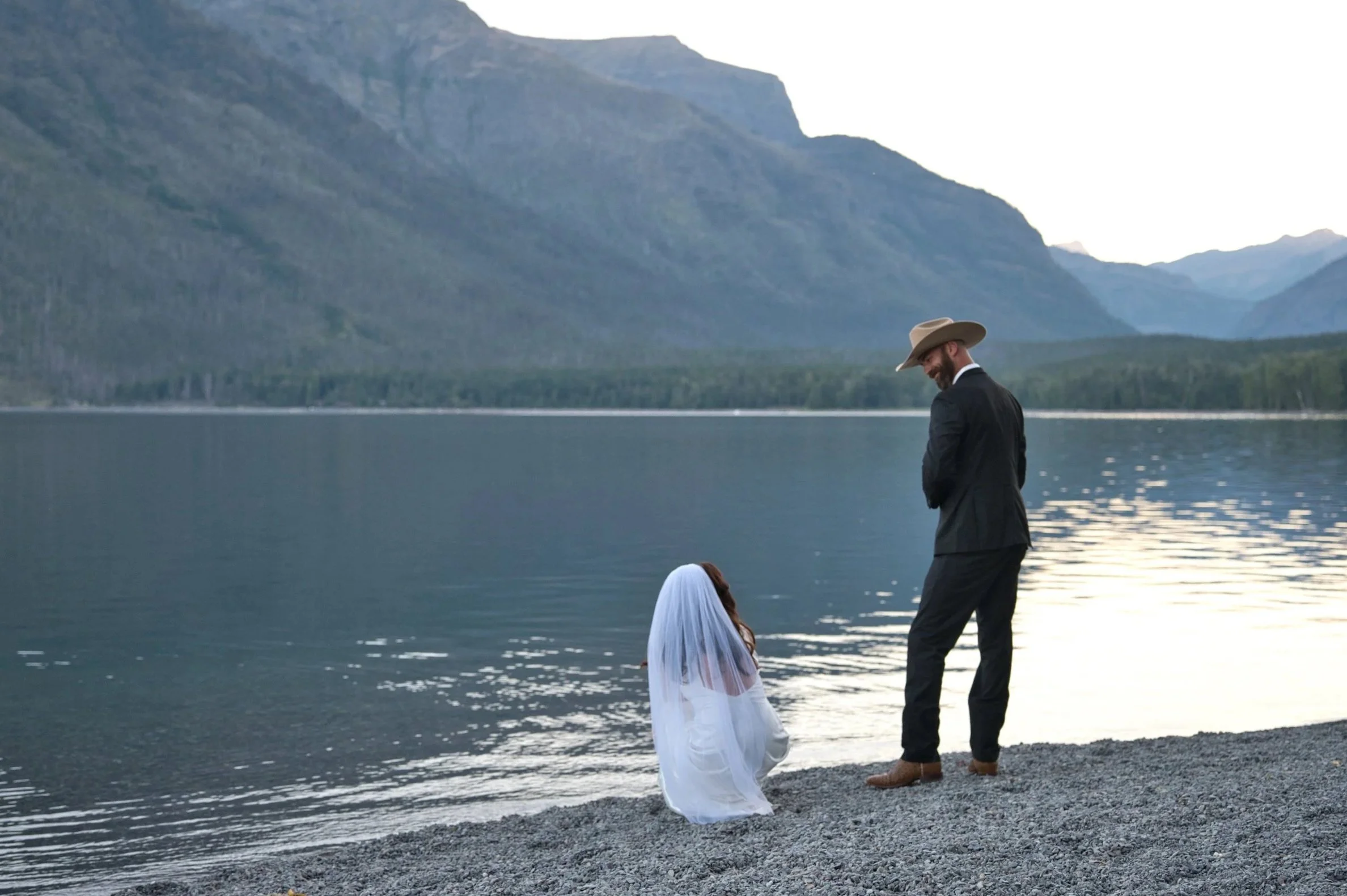 A man in a suit with a cowboy hat standing on a rocky shoreline, looking down at a woman in a white dress with a veil, who is crouching near the water's edge. They are by a lake with mountains in the background during sunset or sunrise.