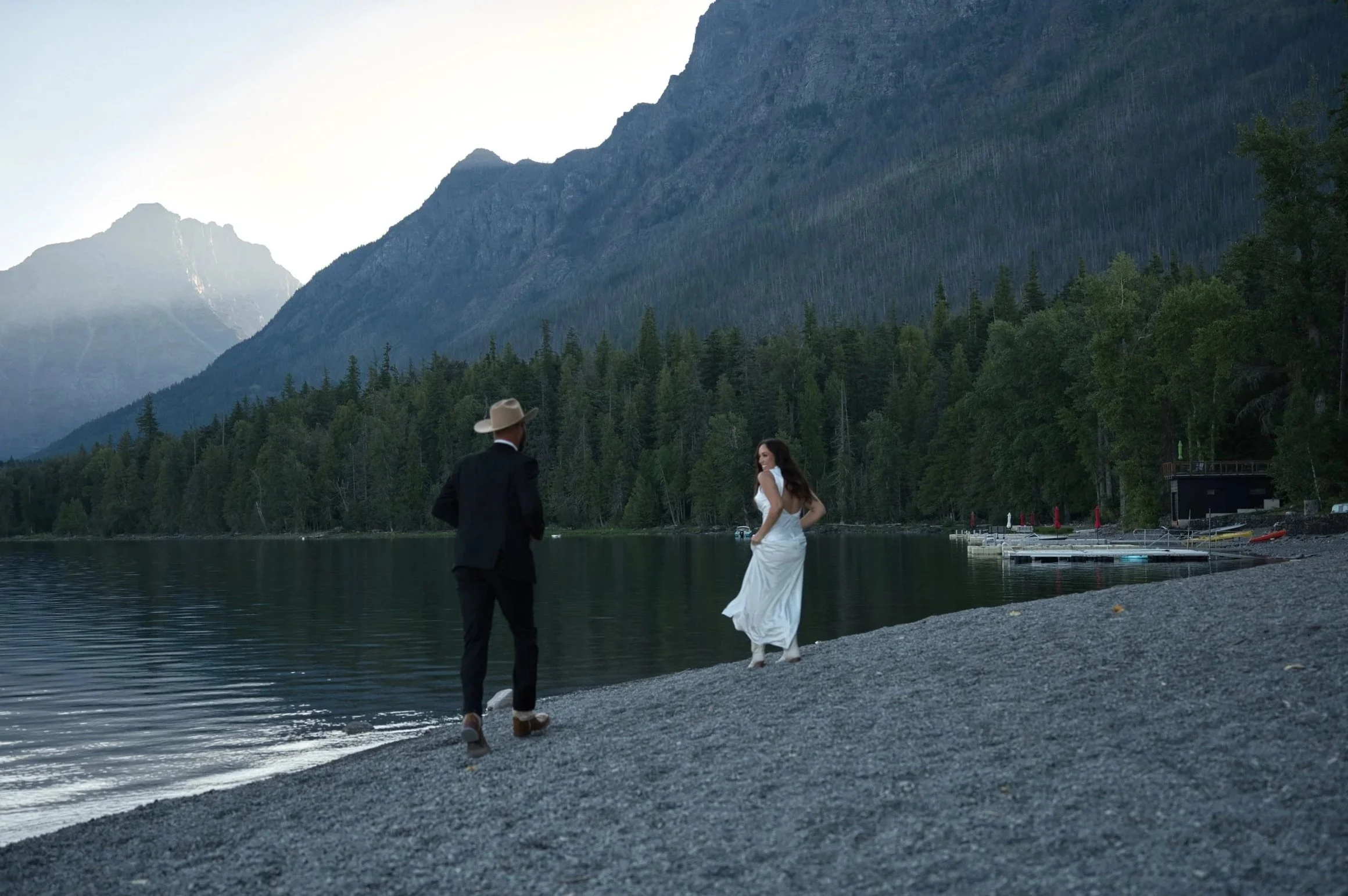 A man and a woman dressed in formal clothes standing on a shoreline by a lake, with mountains and trees in the background.
