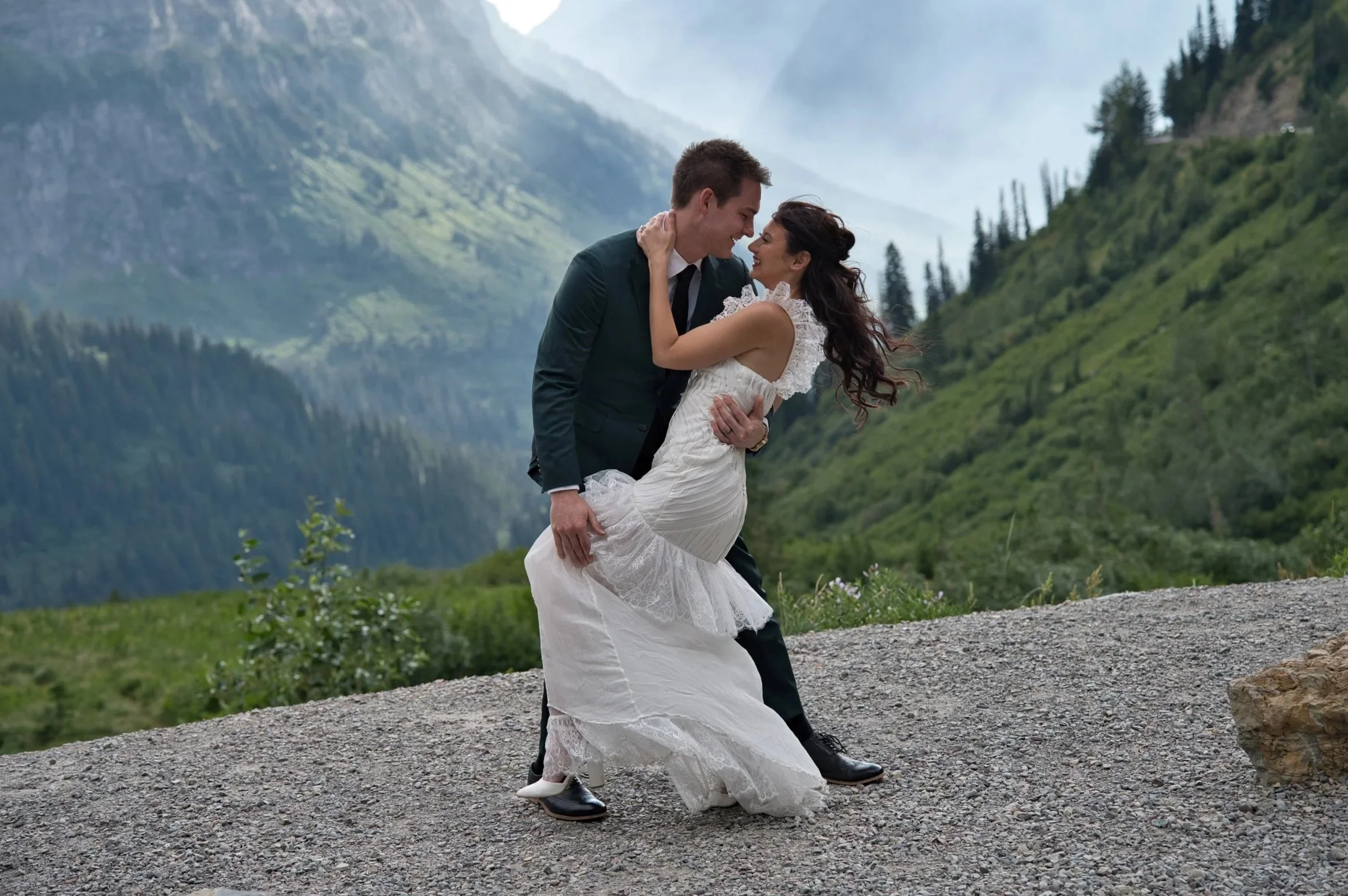 A couple in wedding attire sharing a romantic moment in a scenic mountain landscape, with the man holding the woman in his arms.