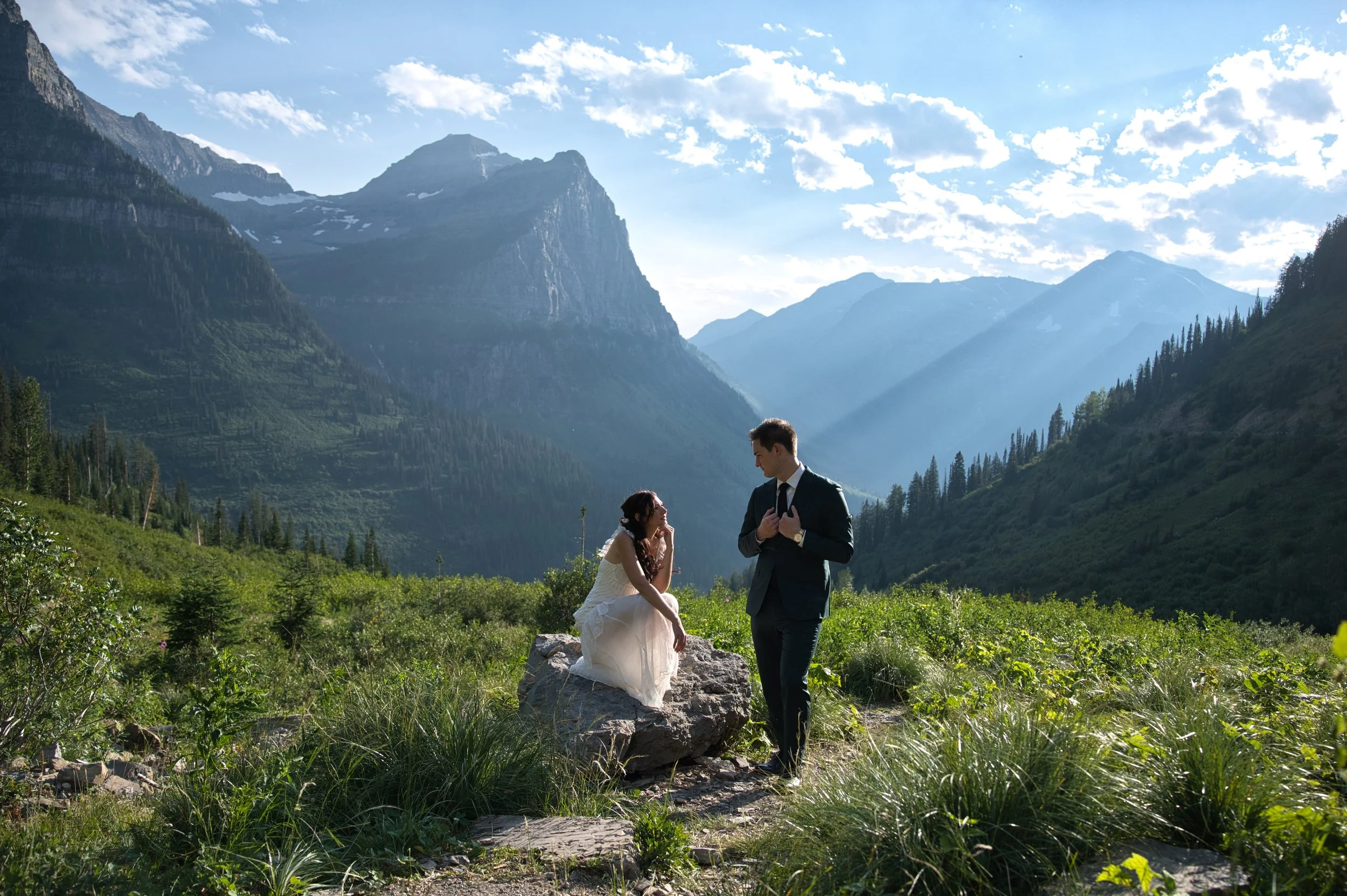 A bride and groom in wedding attire in a lush green valley surrounded by mountains under a partly cloudy sky.