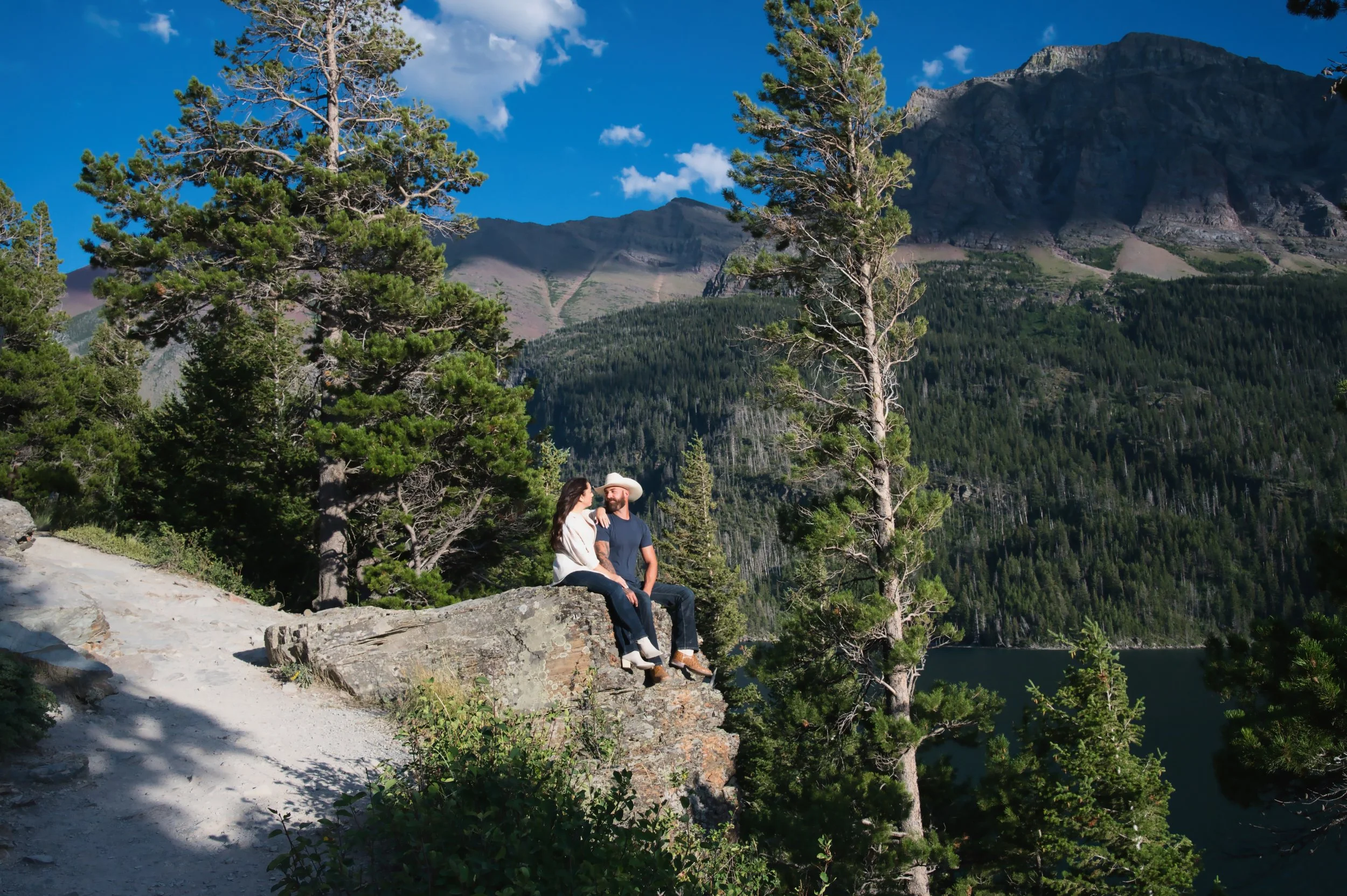 A couple sitting on a large rock in a mountain forest, overlooking a lake with mountain peaks in the background on a sunny day.