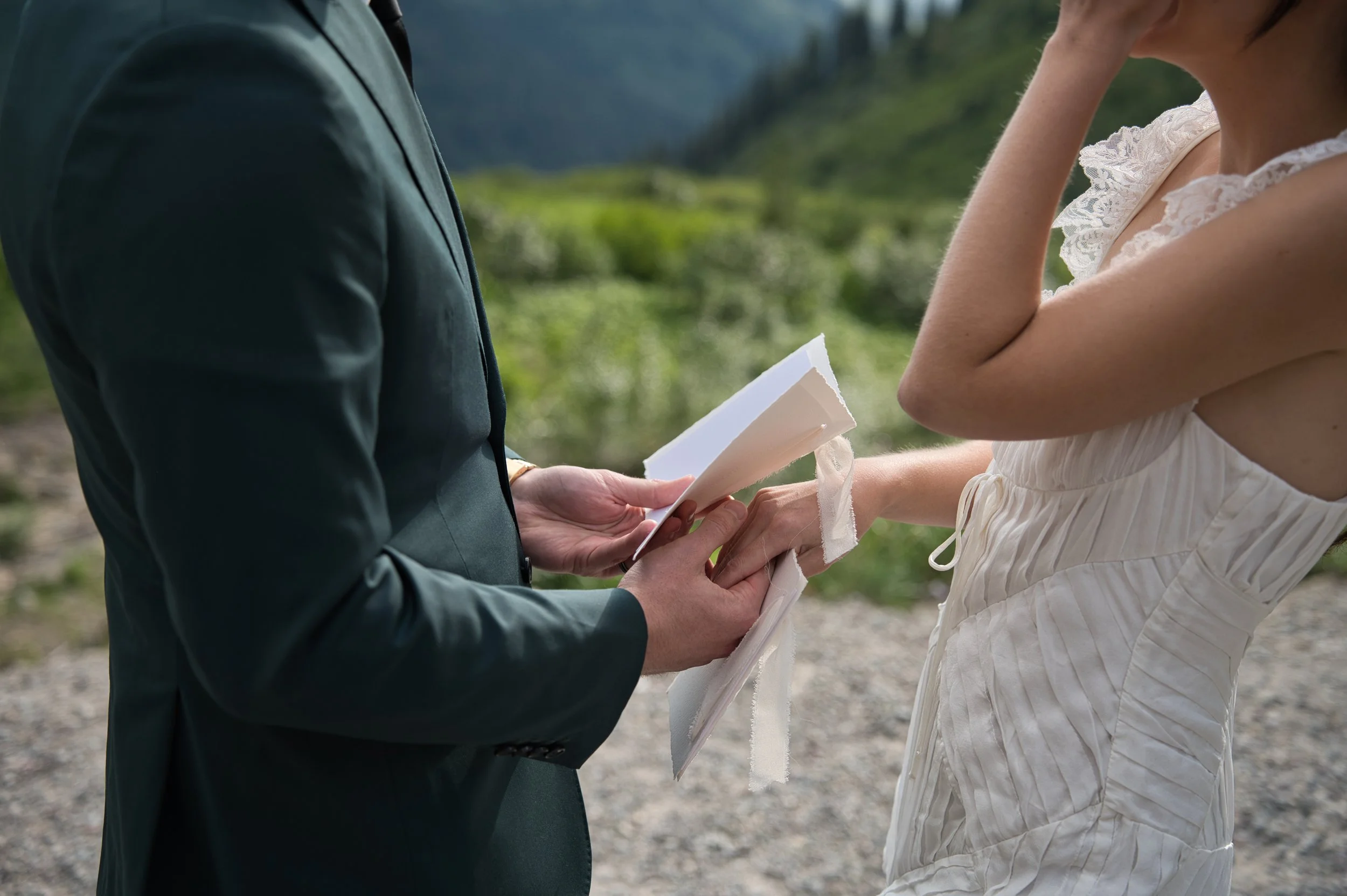 A man and a woman exchanging vows during an outdoor wedding ceremony, with the man holding a small book or card and the woman standing in a light-colored dress.