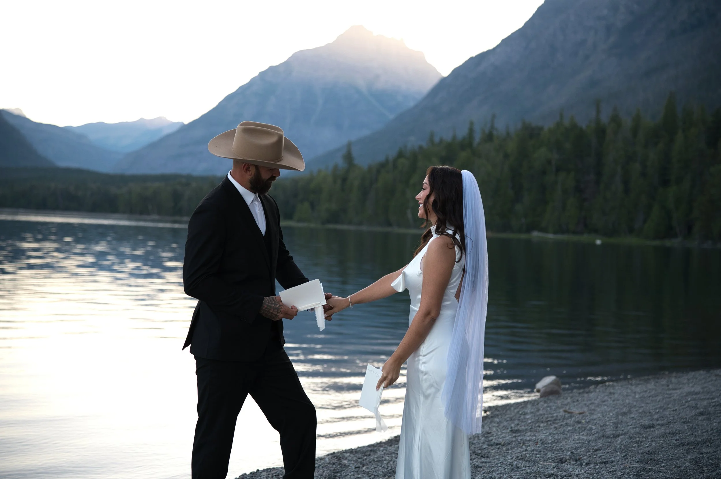 A couple gets married outdoors by a lake with mountains in the background, the groom in a black suit and cowboy hat, the bride in a white dress with a veil, holding hands and smiling at each other.