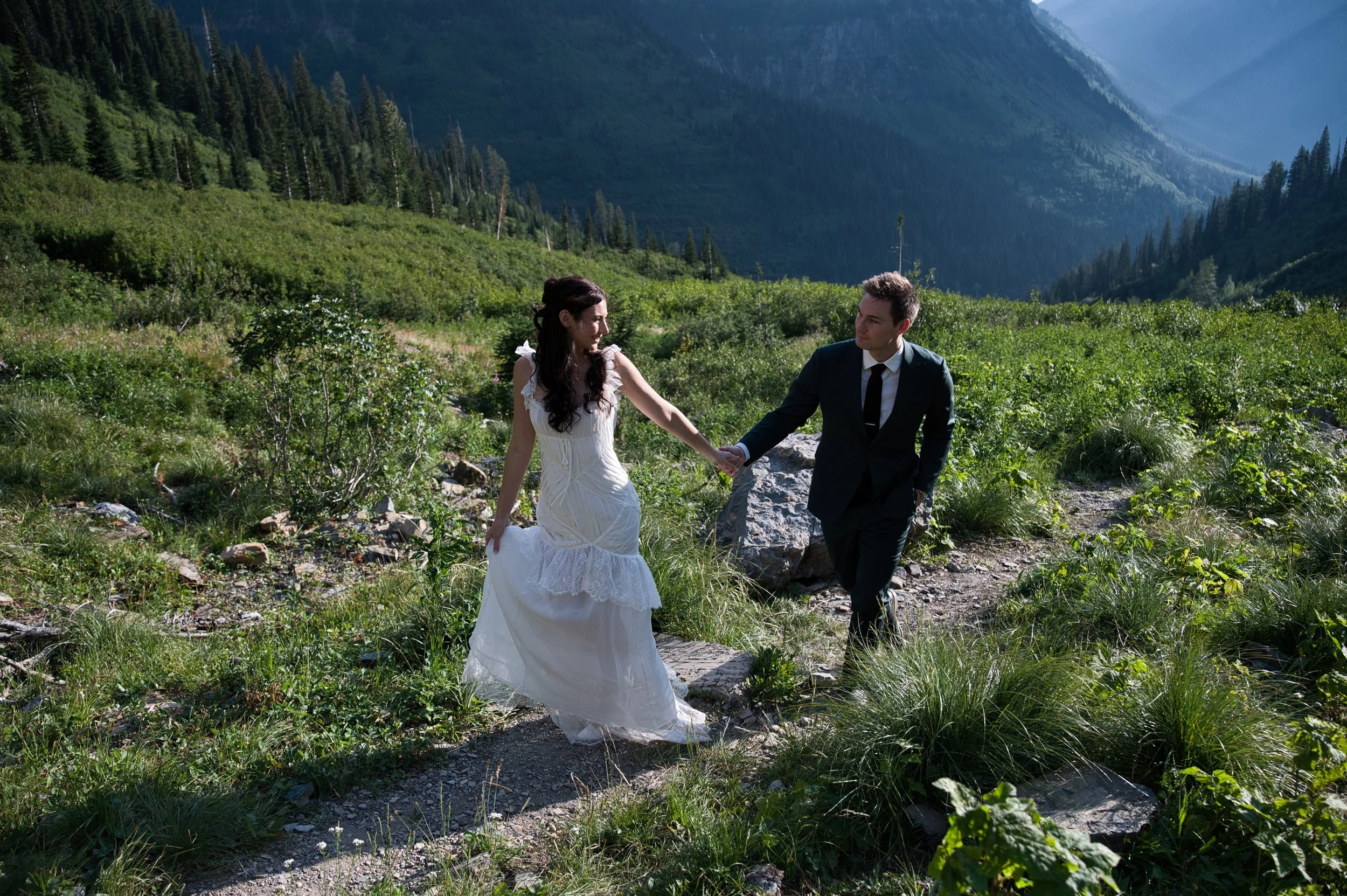 A newlywed couple holding hands and walking through a lush green mountain landscape with tall trees and mountains in the background.