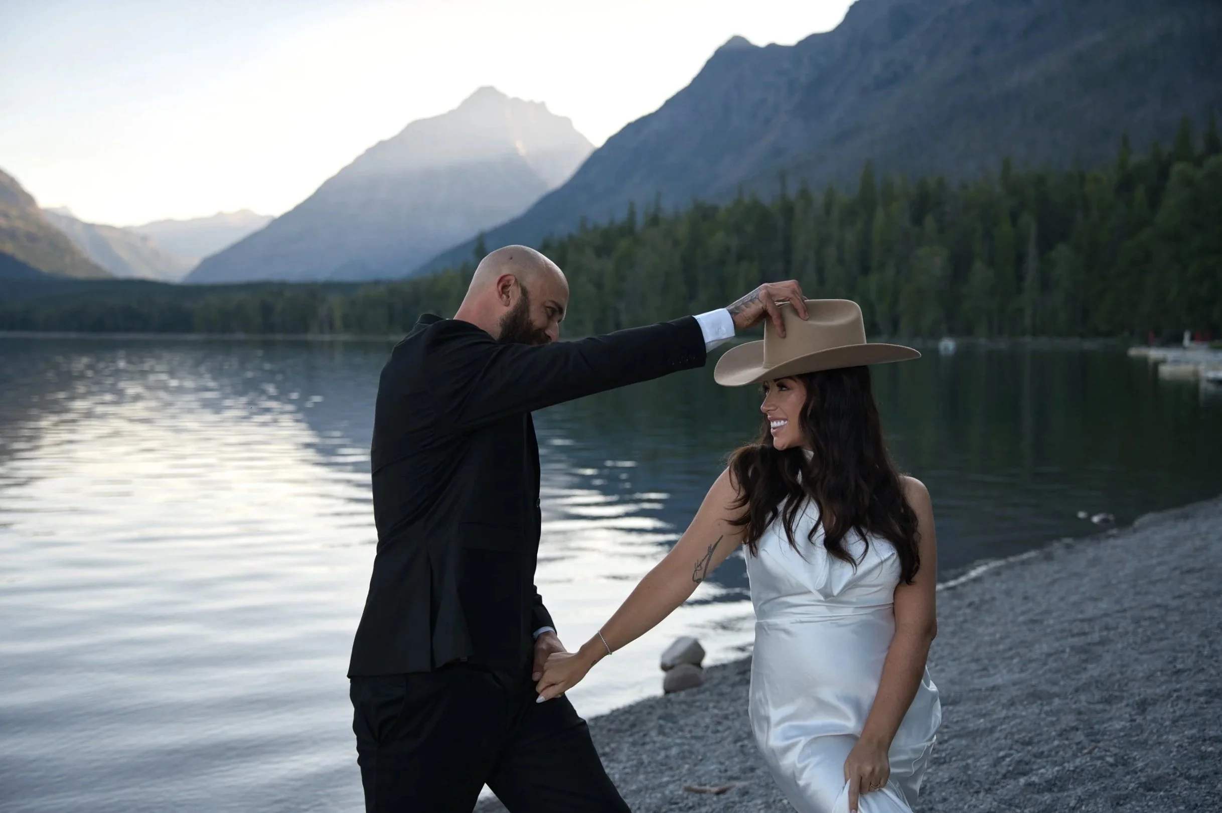 A couple by a lake with mountains in the background, the man is placing a beige wide-brimmed hat on the woman's head, both are smiling; the woman is wearing a white dress and has long dark hair, the man is in a black suit, during sunset or early evening.
