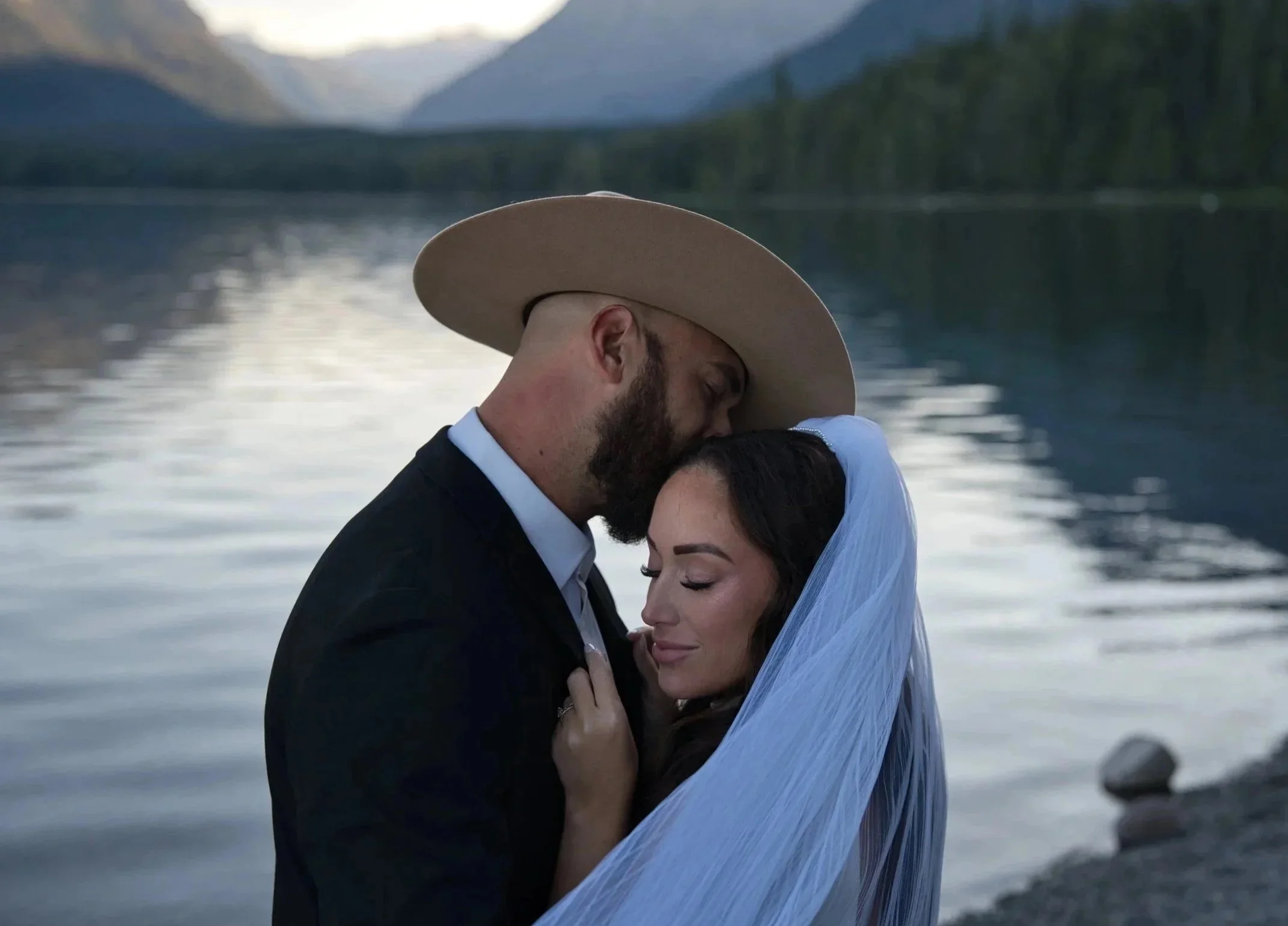A couple embraces by a lake with mountains in the background, the man wearing a large hat and suit, the woman in a white veil with her eyes closed.