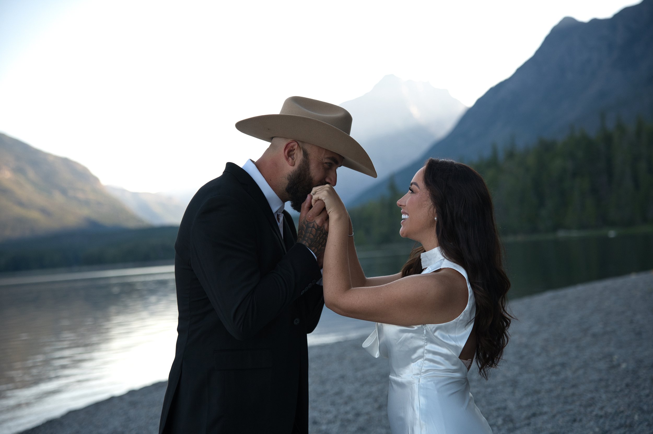 A couple stands by a lake surrounded by mountains. The man, wearing a cowboy hat, is holding hands and kissing the woman's hands, who is smiling at him.