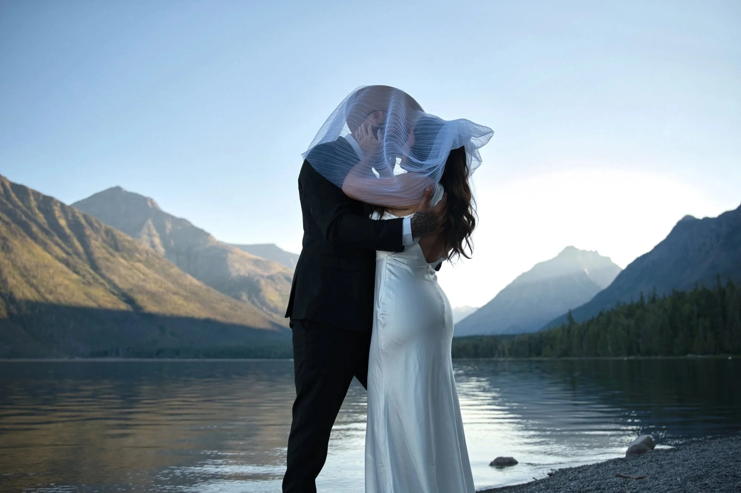 A couple in wedding attire sharing a kiss by a lake with mountains in the background, and the bride wearing a veil.