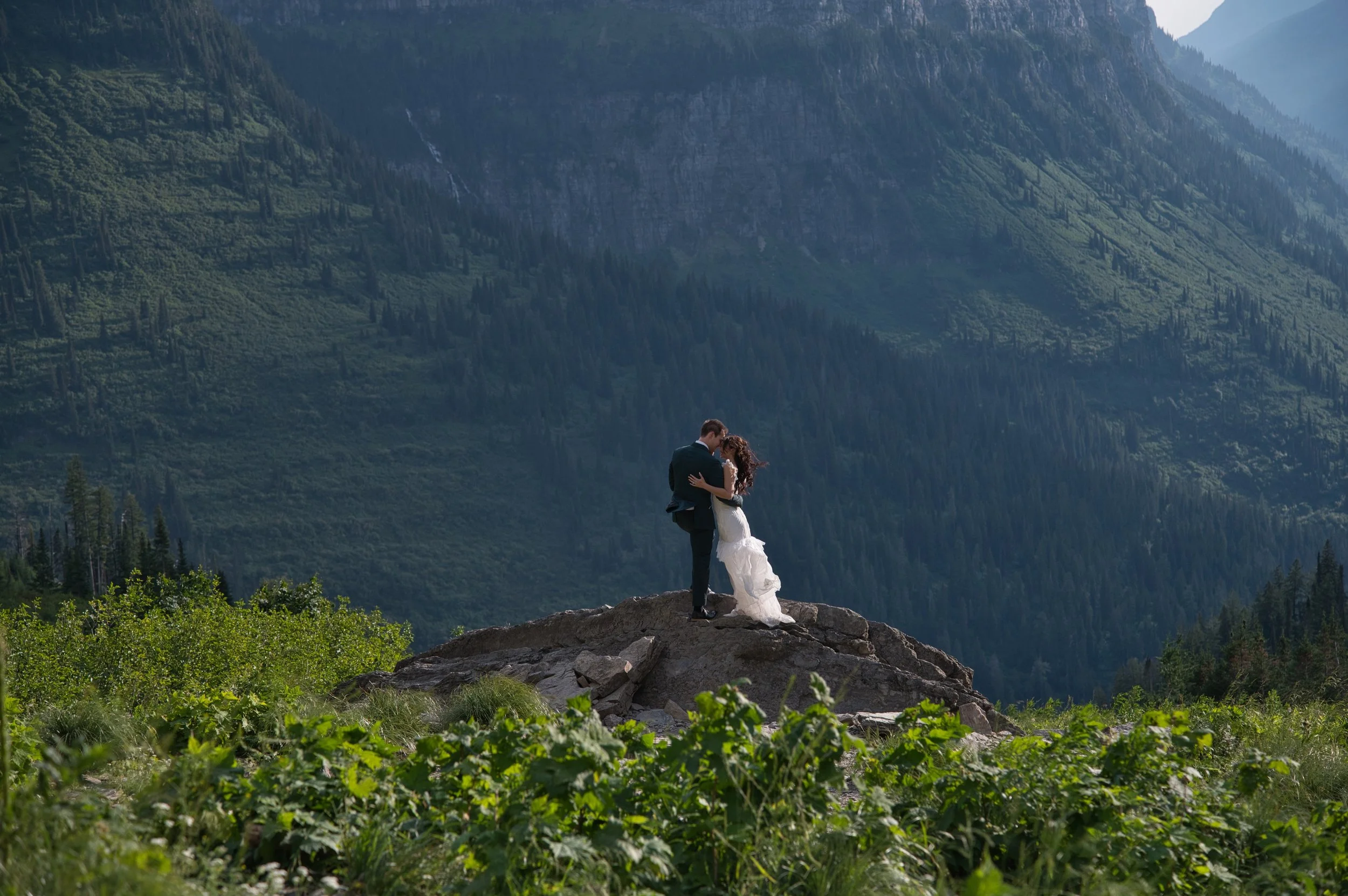 A couple in wedding attire standing on a rock, embracing in a mountain landscape with lush greenery and tall trees.