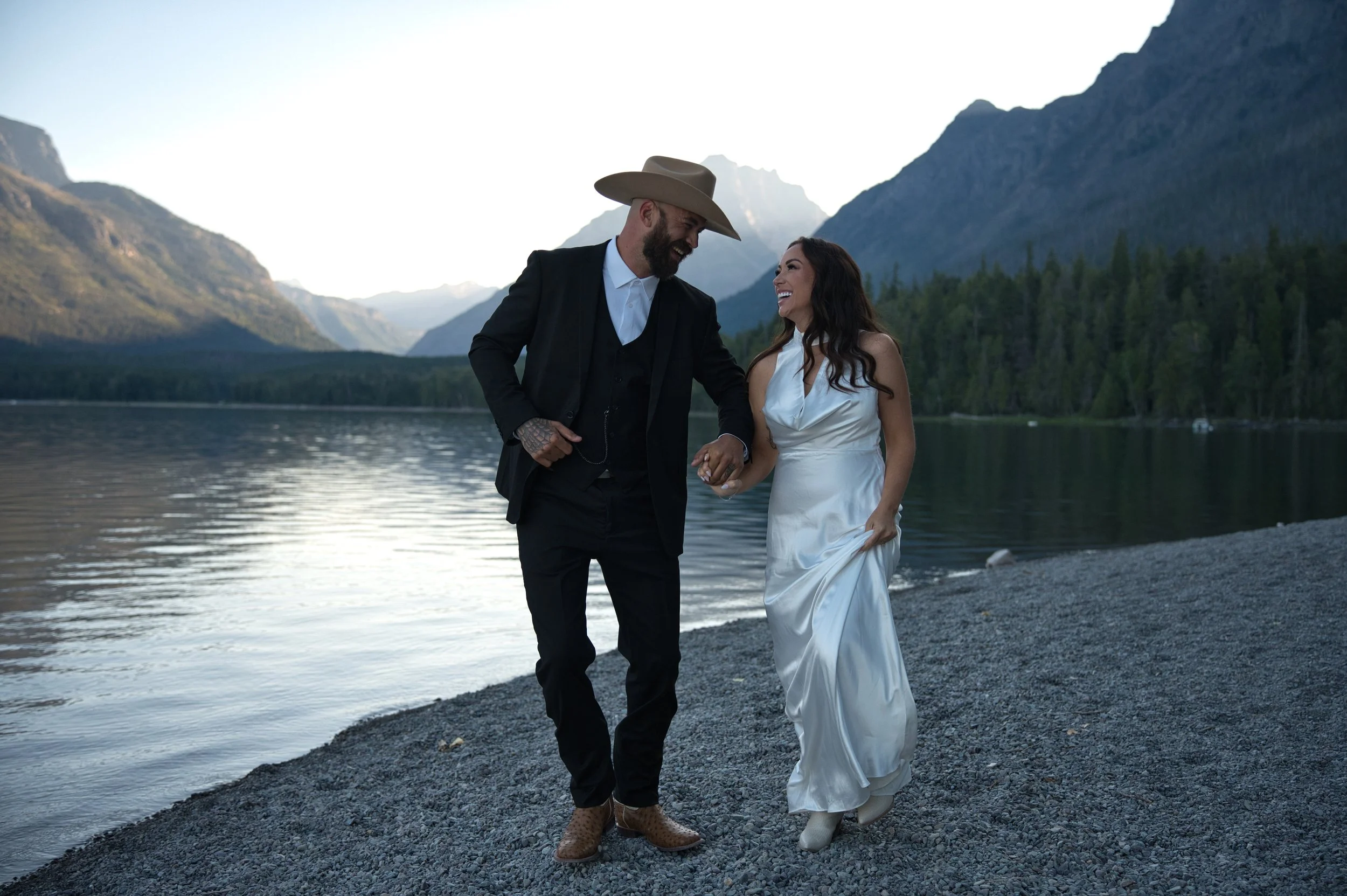 A couple dressed in wedding attire walking hand in hand along a rocky lakeshore with mountains and trees in the background.