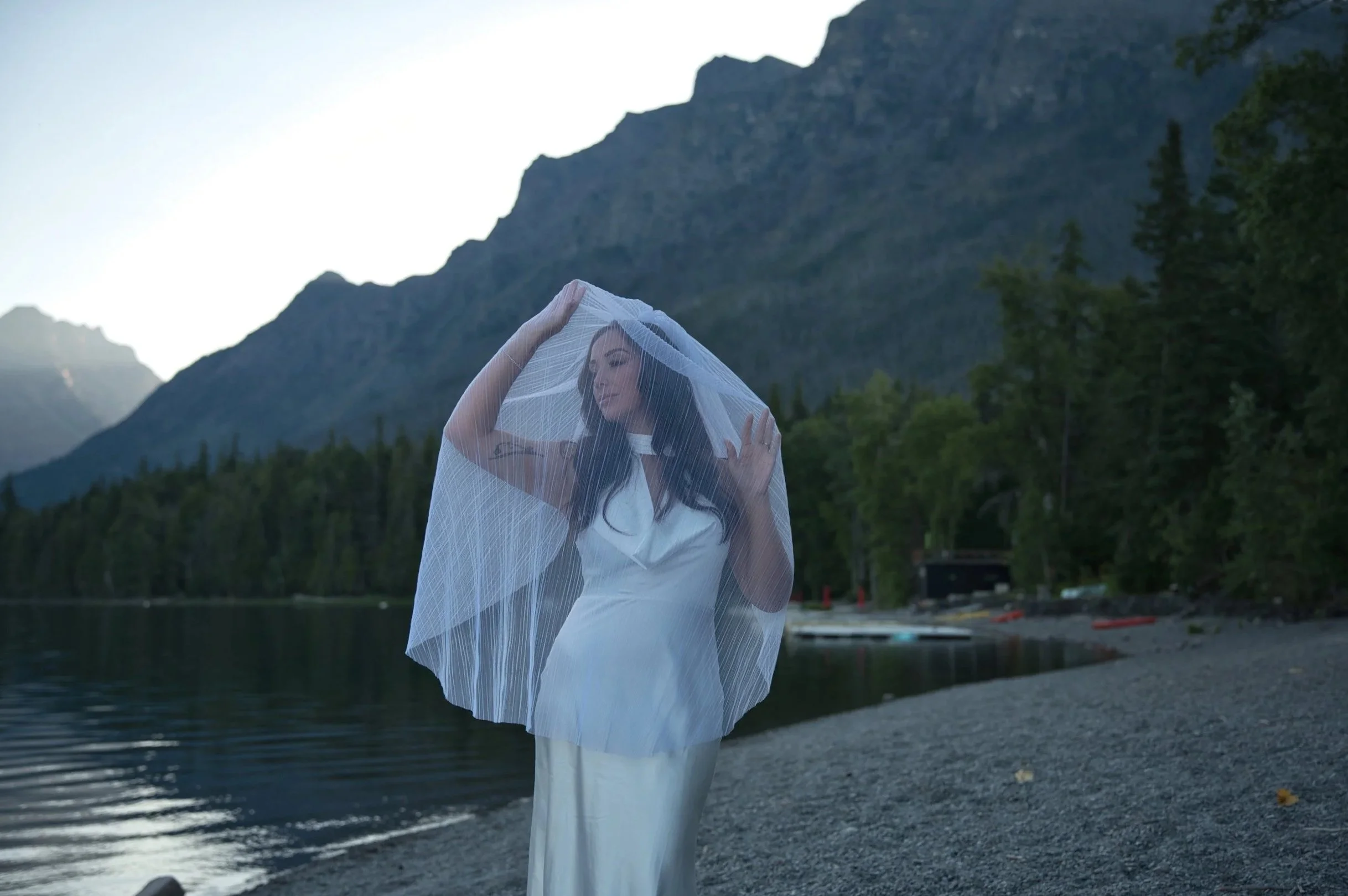 Woman in a white dress with a veil standing on a beach near a lake with mountains and trees in the background.