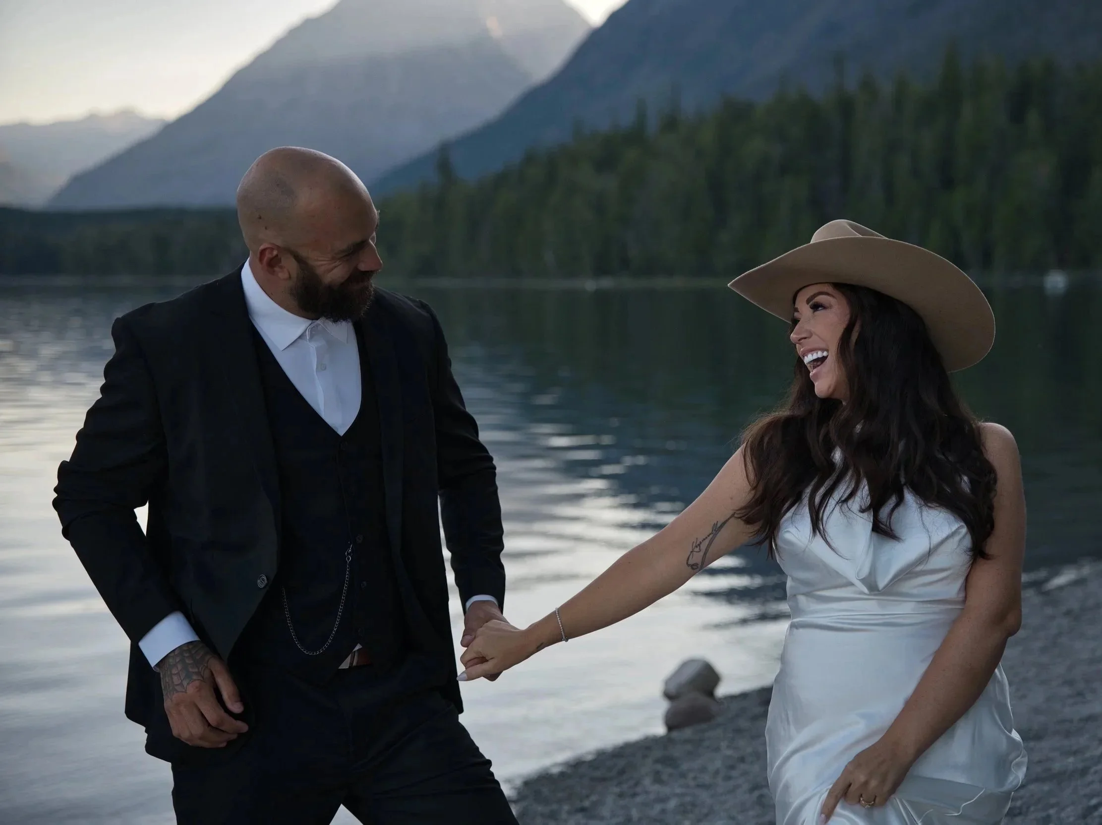 A man and woman holding hands near a lake with mountains in the background, smiling at each other.