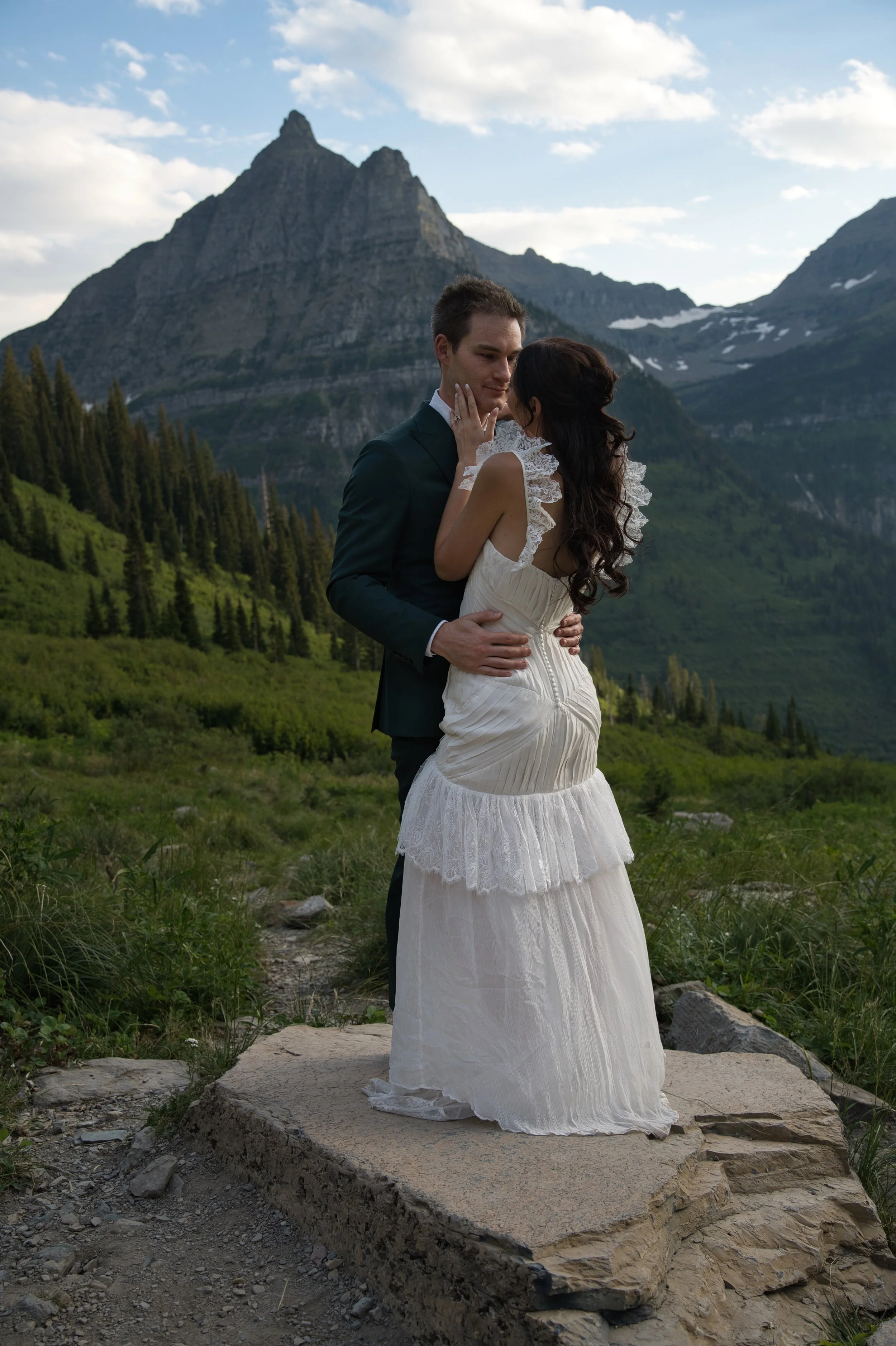 A couple in wedding attire standing on a rock in a mountainous landscape with green foliage and snow-capped peaks in the background.
