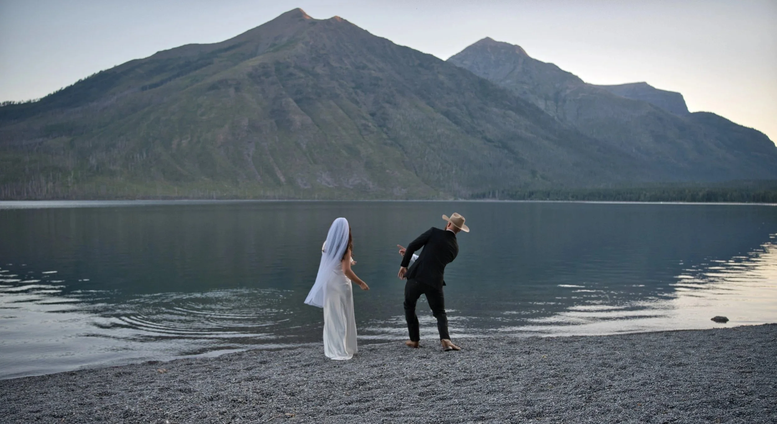 A bride and groom walking along the shoreline of a lake with mountains in the background at sunset.