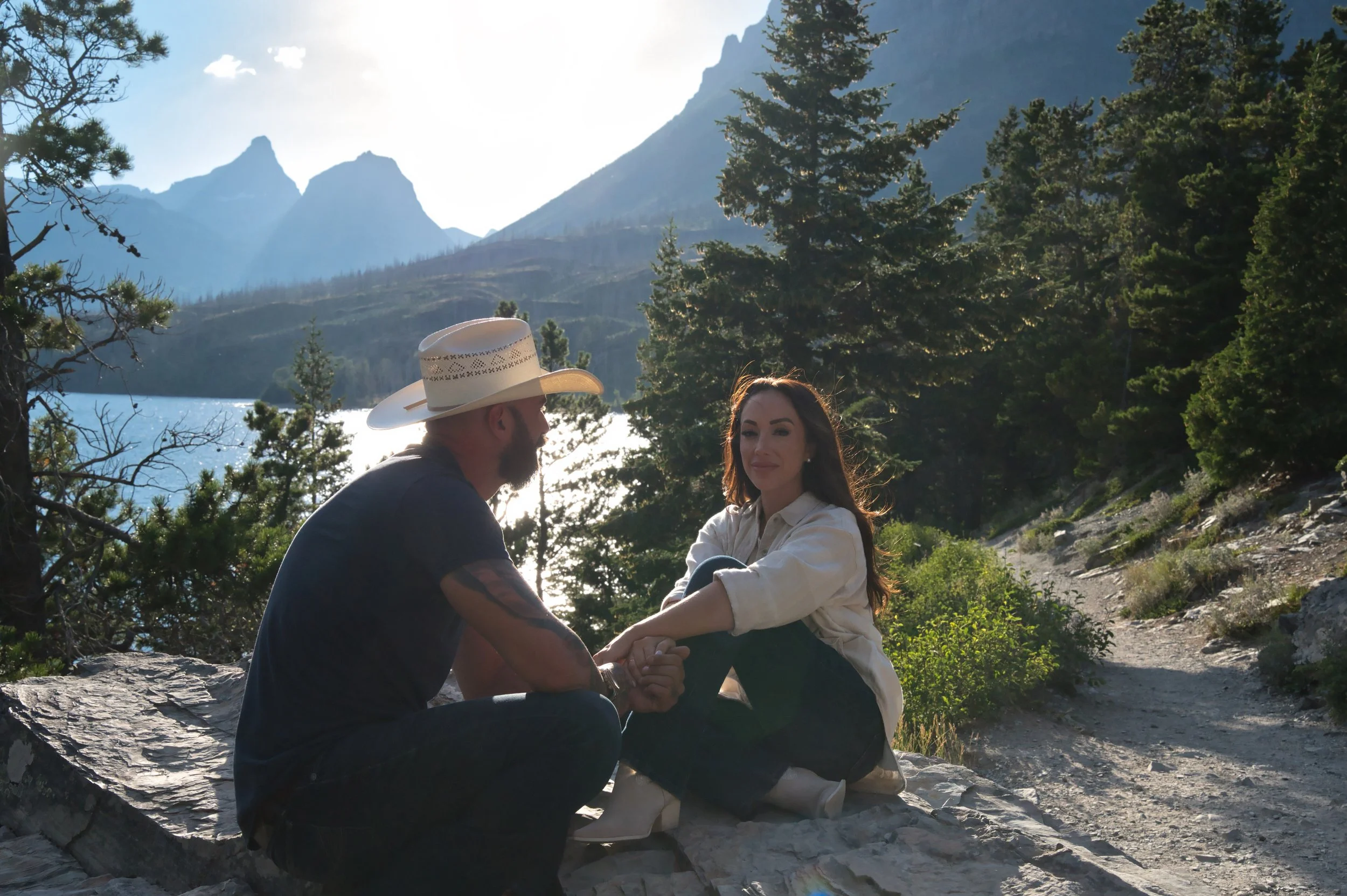 A man and woman sitting on rocks outdoors, holding hands and looking at each other, with a lake, mountains, and trees in the background during daytime.