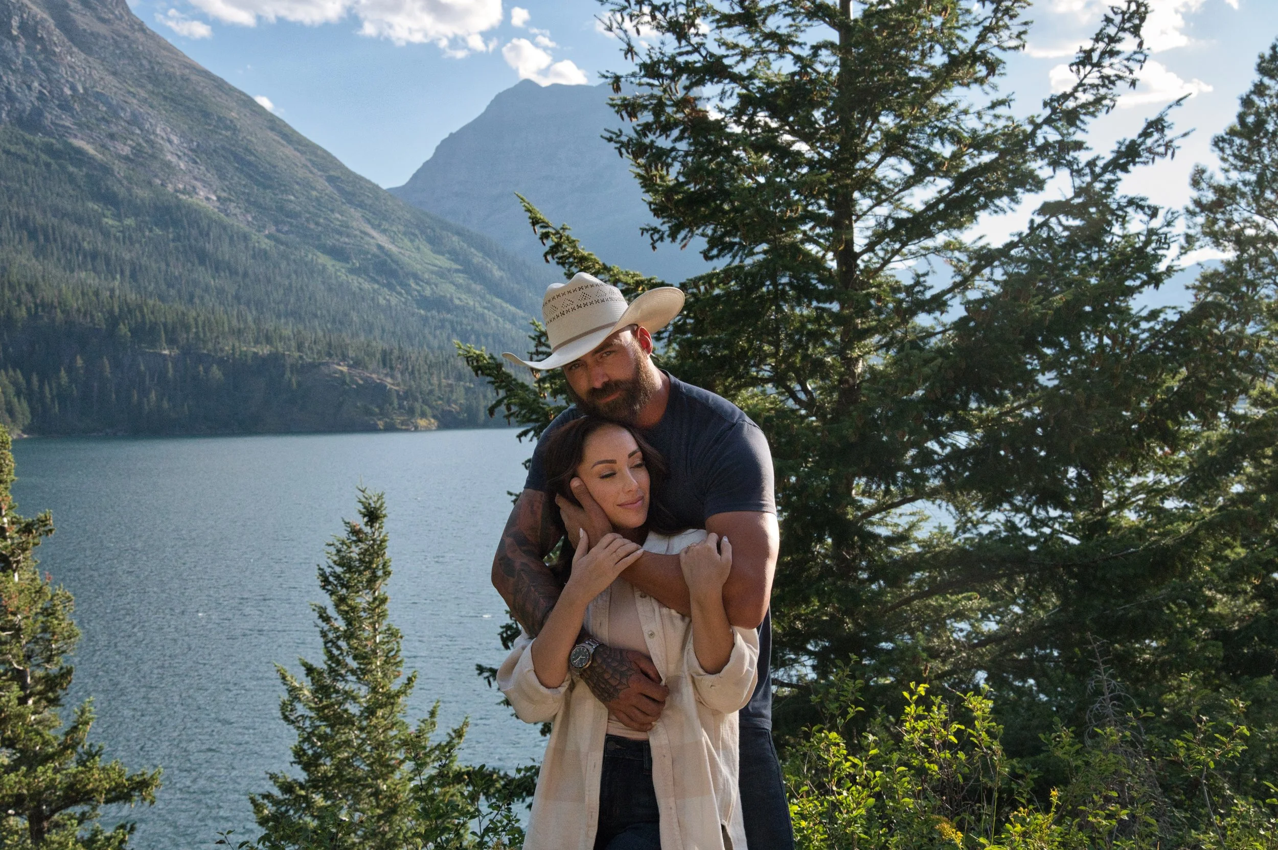 A man and woman embracing near a lake with mountains and trees in the background, during daytime.