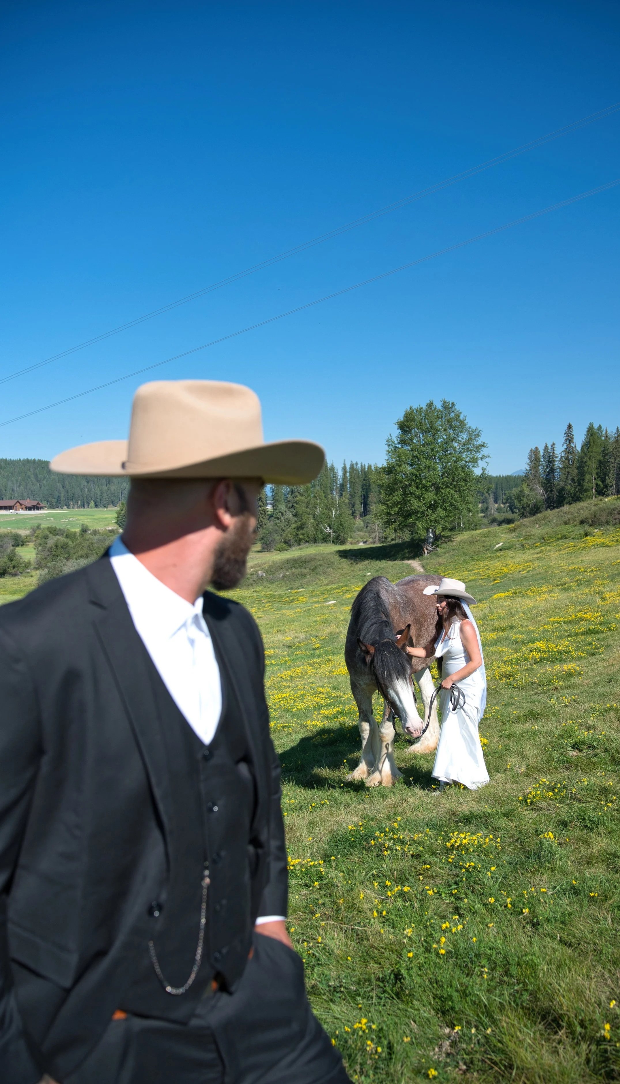 A man in a black suit and beige cowboy hat watching a woman in a white dress and cowboy hat pet a large brown and white horse in a green field with yellow flowers under a clear blue sky.