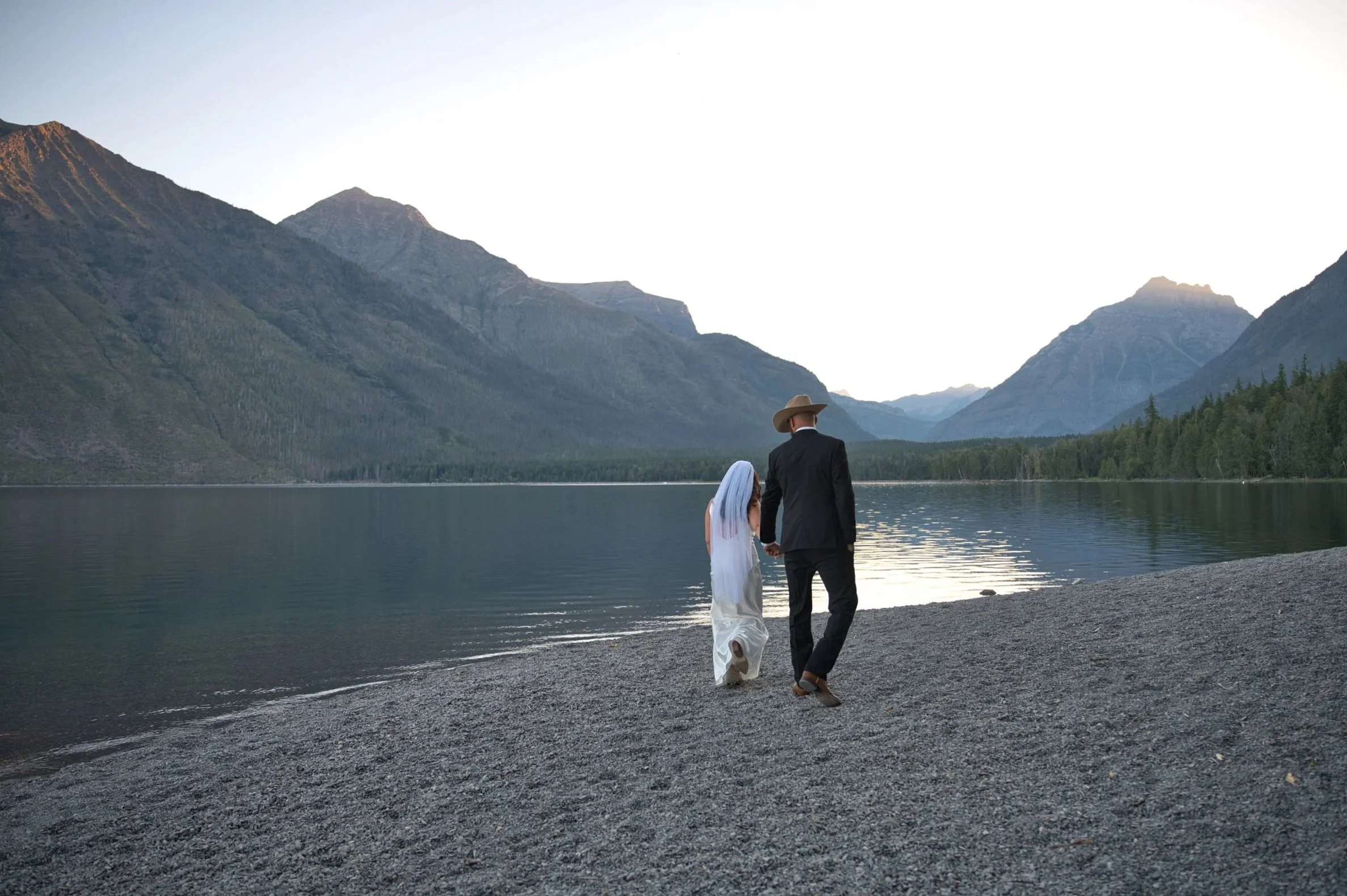 A couple dressed in wedding attire walking hand in hand along the shore of a lake surrounded by mountains at sunset.