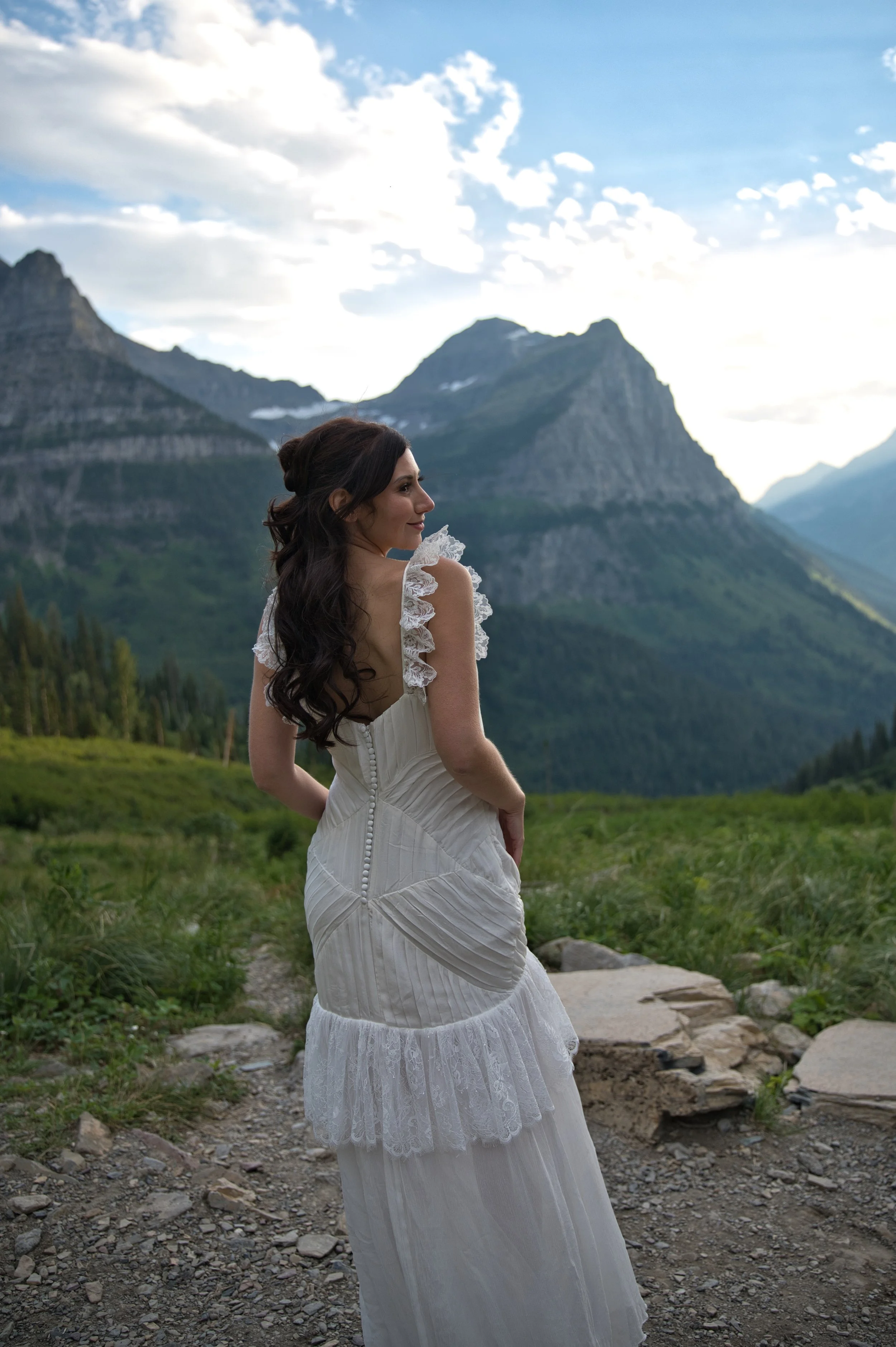 A woman in a white dress with lace details standing outdoors on a mountain trail, with mountains and a cloudy sky in the background.