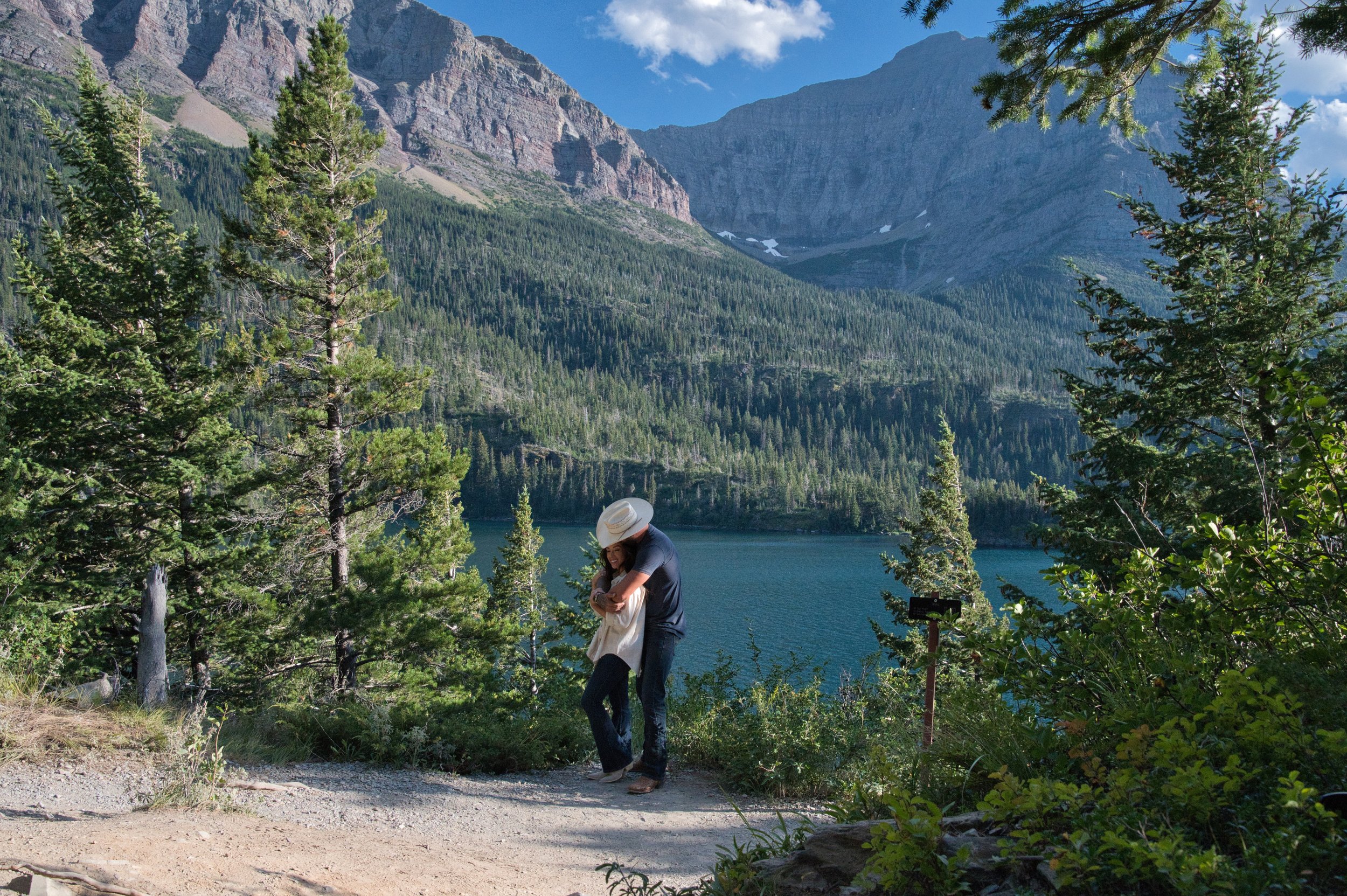 Couple embracing on a trail near a lake surrounded by pine trees and mountains under a blue sky.