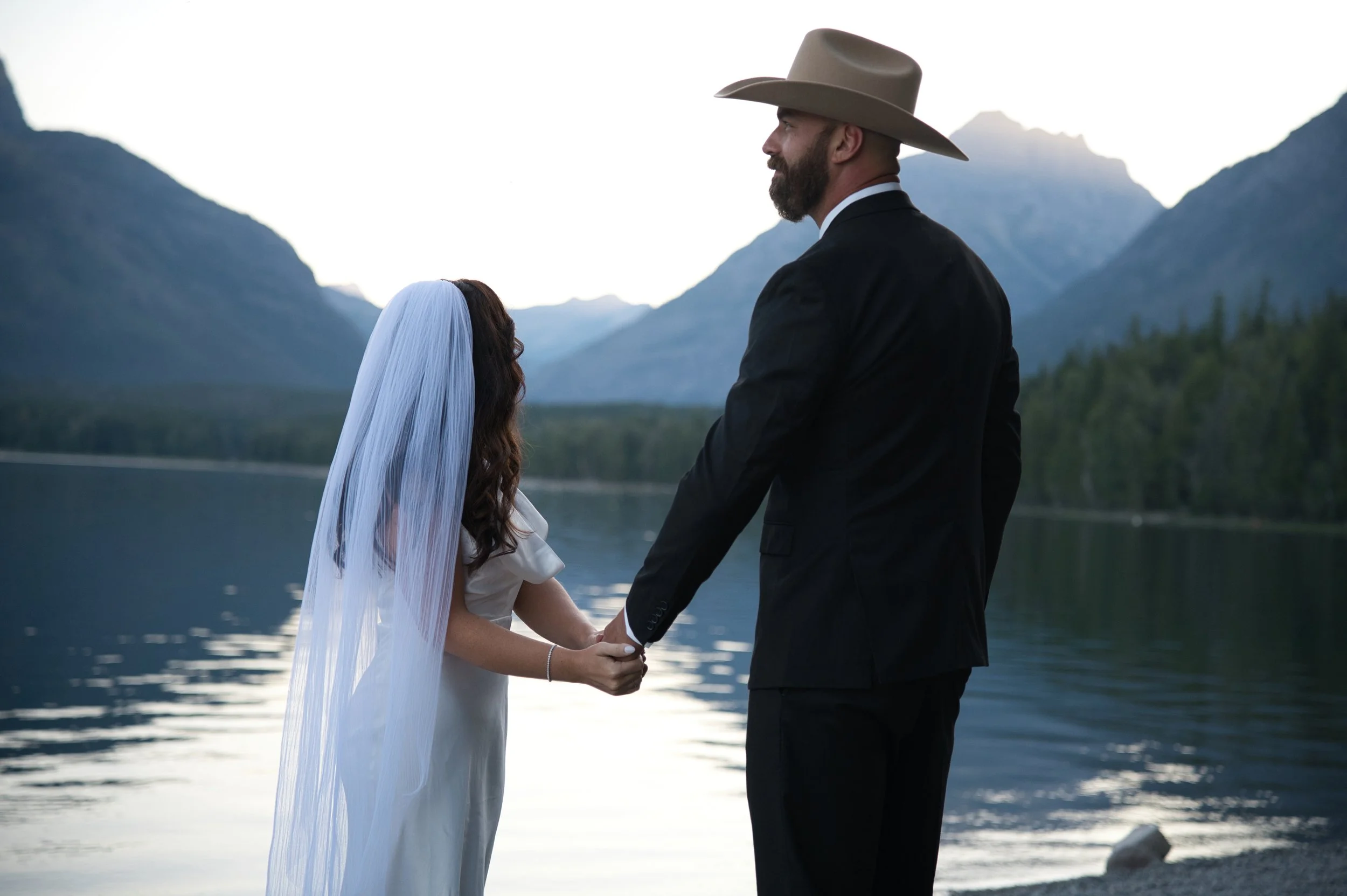 A couple holding hands by a lake with mountains in the background during sunset.
