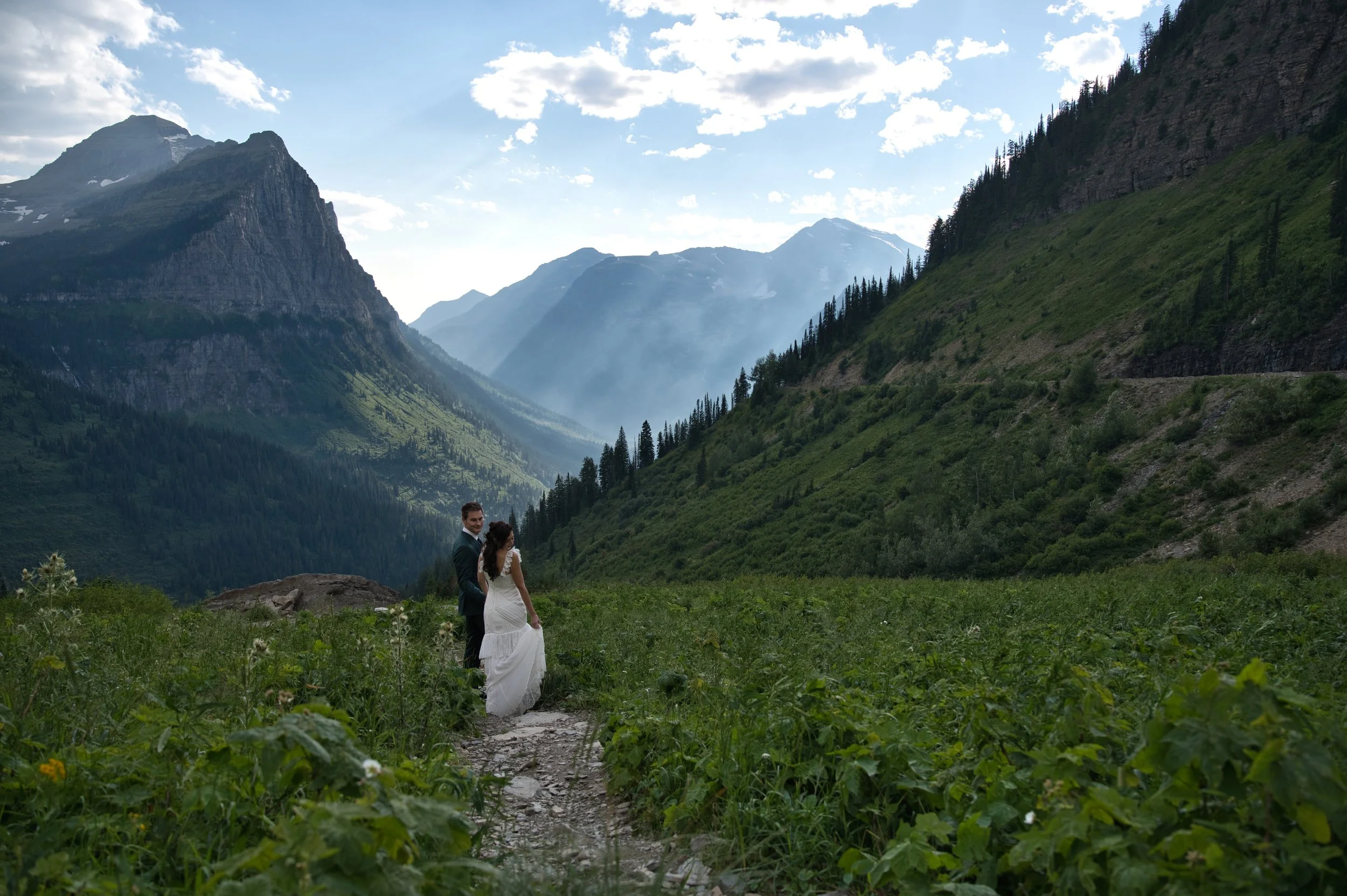 A bride and groom standing on a dirt trail surrounded by green foliage in a mountainous landscape with tall peaks and a partly cloudy sky.
