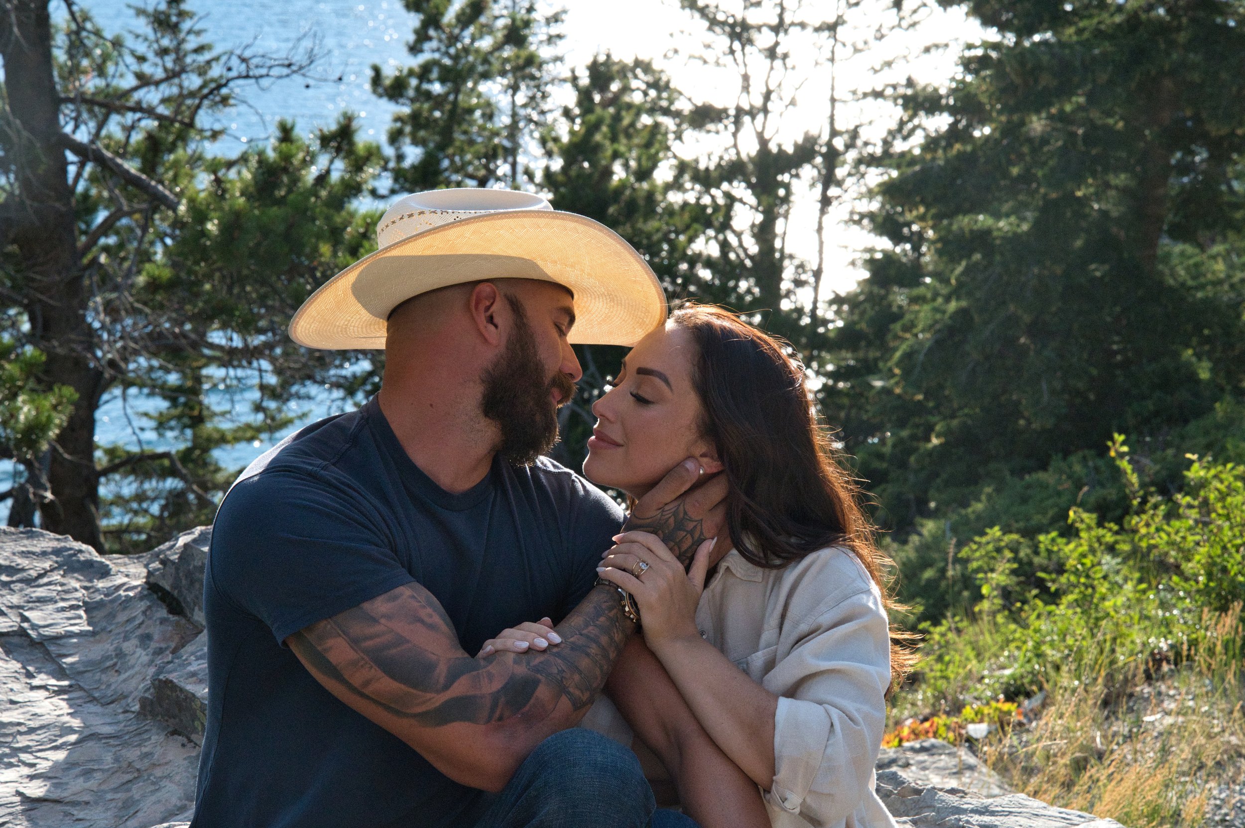 A man and woman sitting outdoors on rocks, looking into each other's eyes, with trees and sunlight in the background.
