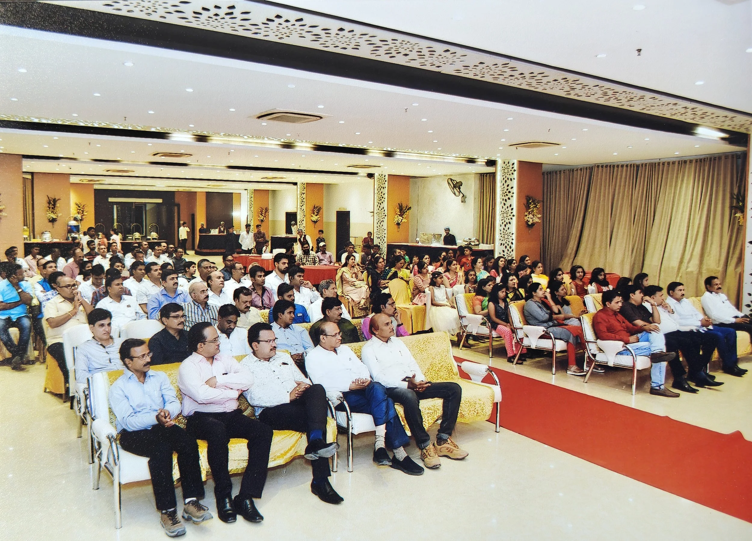 People seated in rows at a formal event in a large, decorated banquet hall with gold and cream-colored decor, floral arrangements on the walls, and a red carpet running through the center.