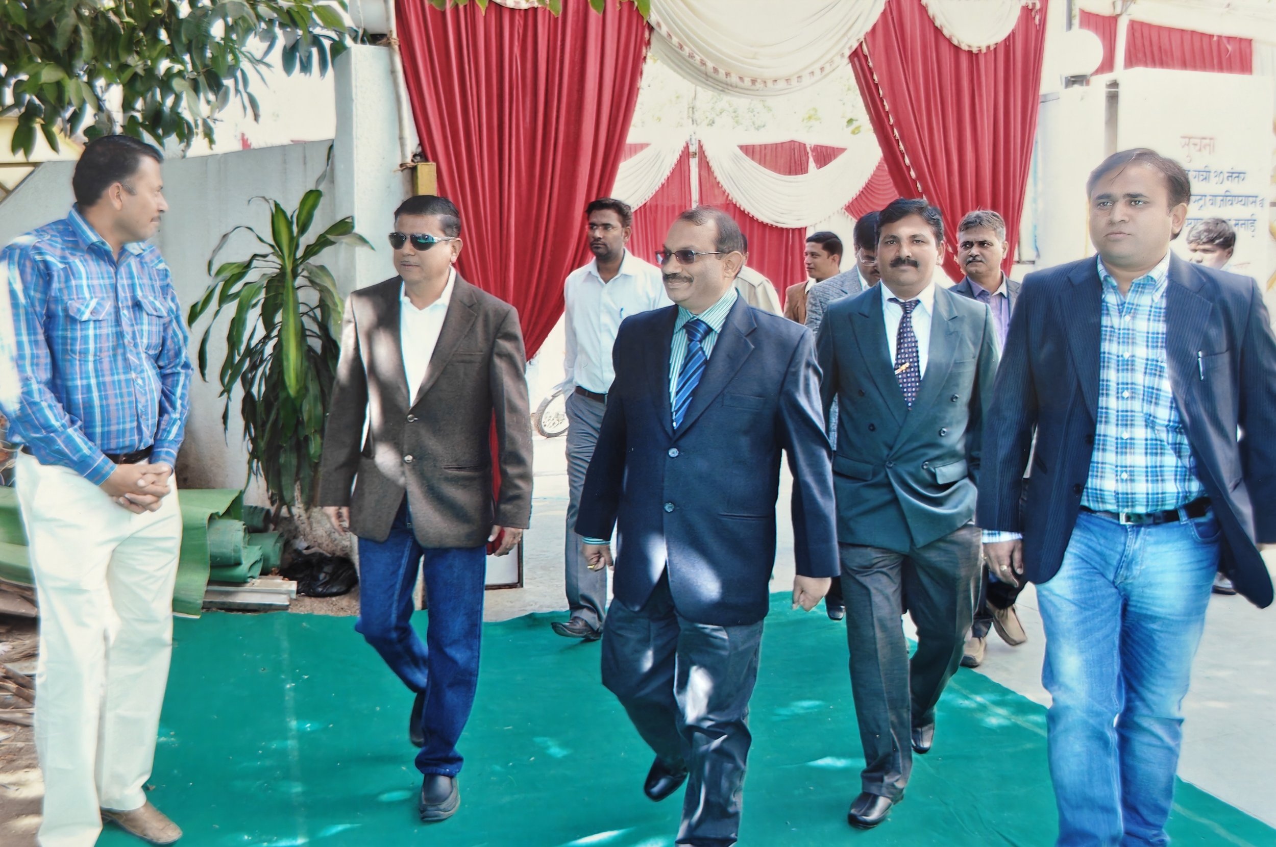 Group of men in formal attire walking on a green carpet outside a decorated venue with red and white drapes.