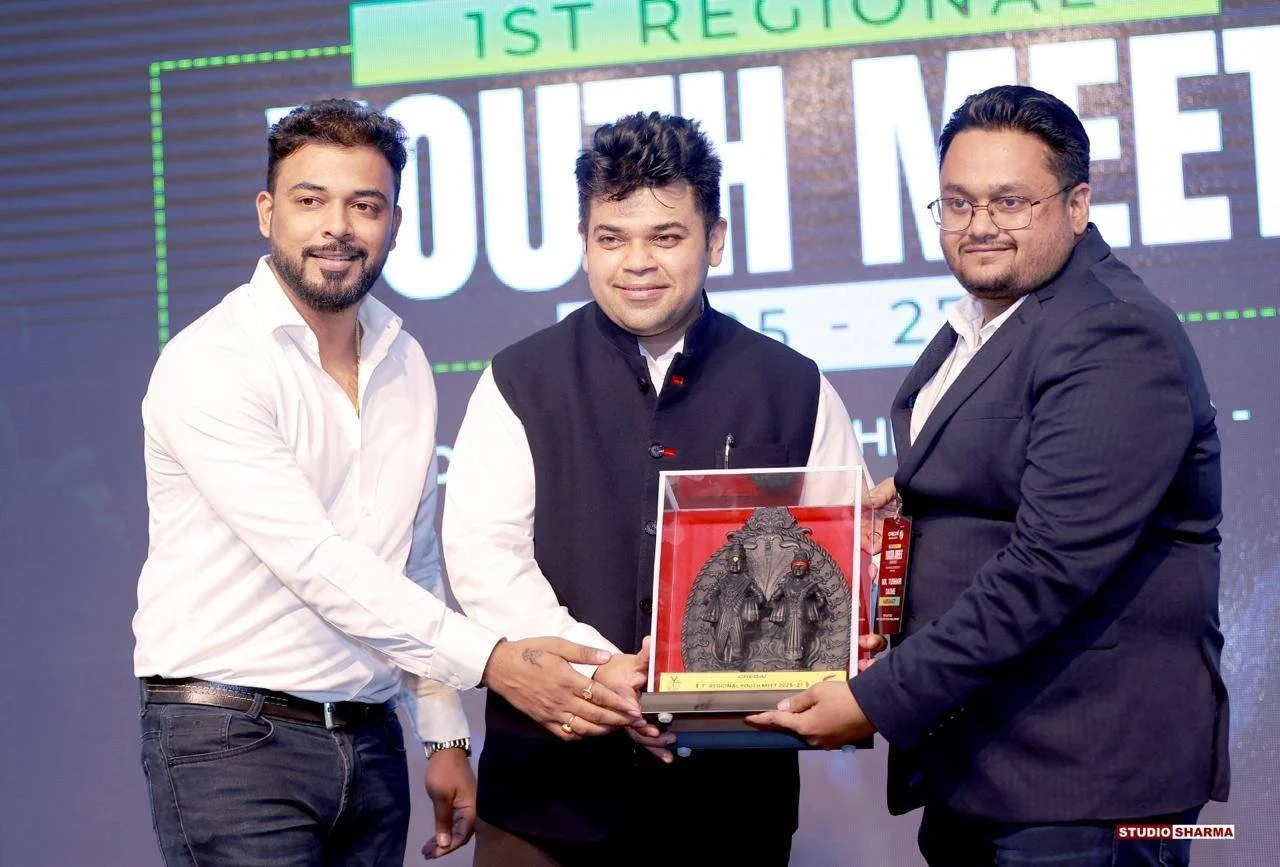 Three men standing together on stage during an award ceremony, holding a glass box containing a statue of two figures. The background displays text reading '1st Regional Youth Mela' and other details.