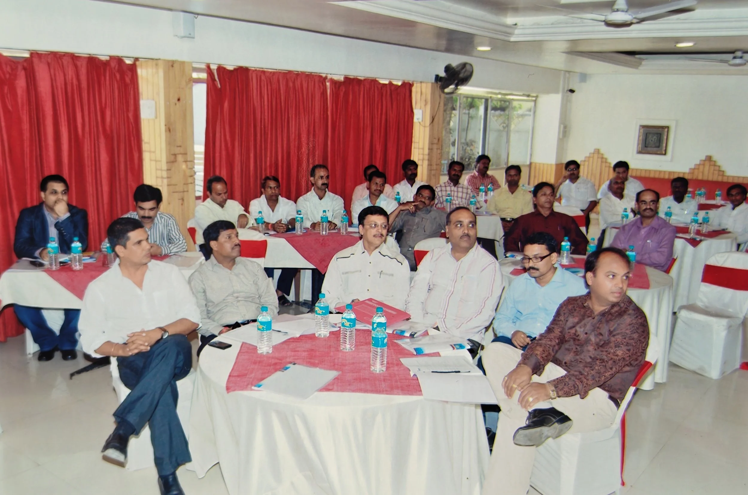 A group of men attending a meeting or conference in a banquet hall with red curtains and tables with water bottles and documents.