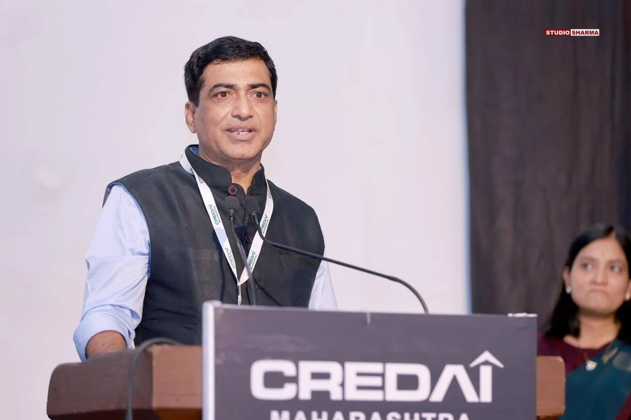 A man in a black vest and blue shirt speaking at a podium with a sign that reads "CREDAI MAHARASHTRA." A woman in a dark dress stands in the background to the right.