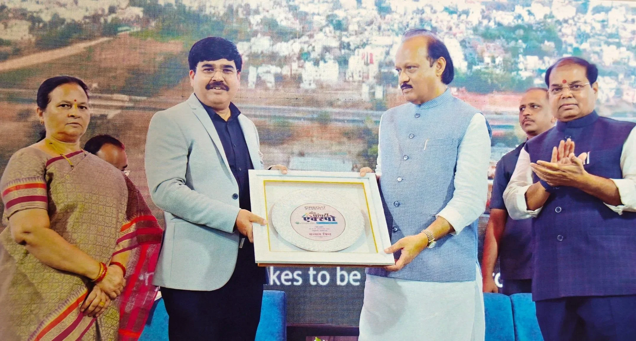 A group of five people on stage during an award ceremony, with two men in the center holding a framed award, surrounded by a woman and three men, with a backdrop of a cityscape.
