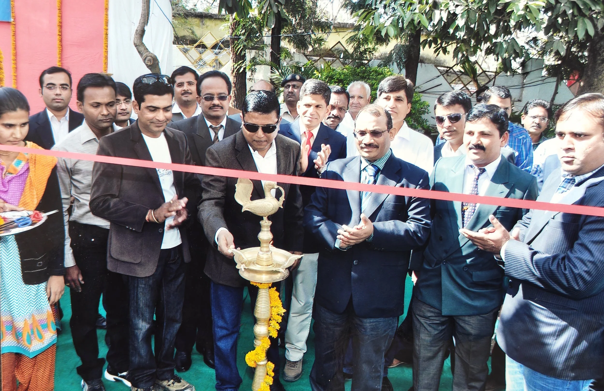 A group of men and women celebrating an inauguration under a red ribbon, with a traditional lamp in the center, outdoors with trees and colorful decorations in the background.