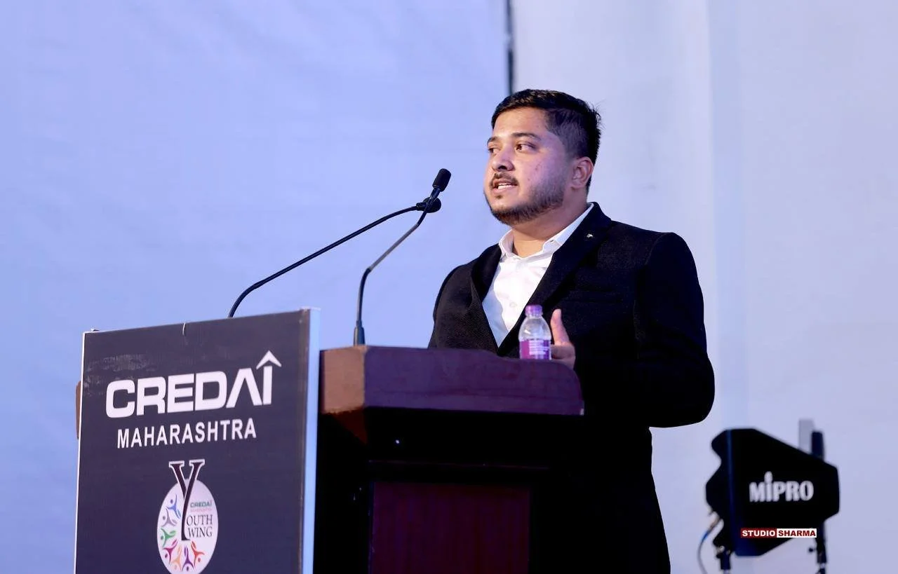 A man in a black suit speaking at a podium during an event, with a sign on the podium reading 'CREDai MAHARASHTRA' and a logo underneath. There is a purple water bottle on the podium and a camera to the right side recording the event.