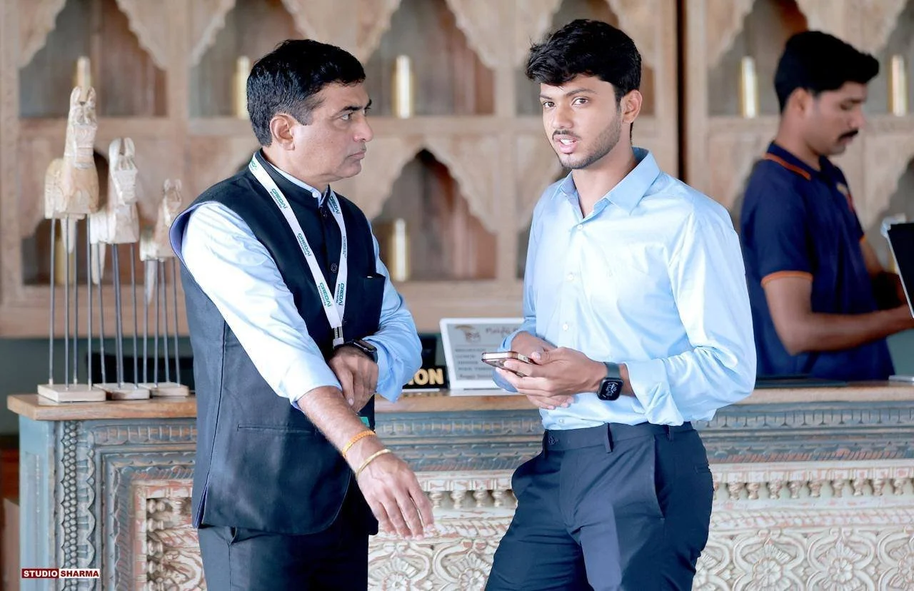 Two men having a conversation in a hotel lobby, with one holding a smartphone, and a staff member working at the reception desk in the background.