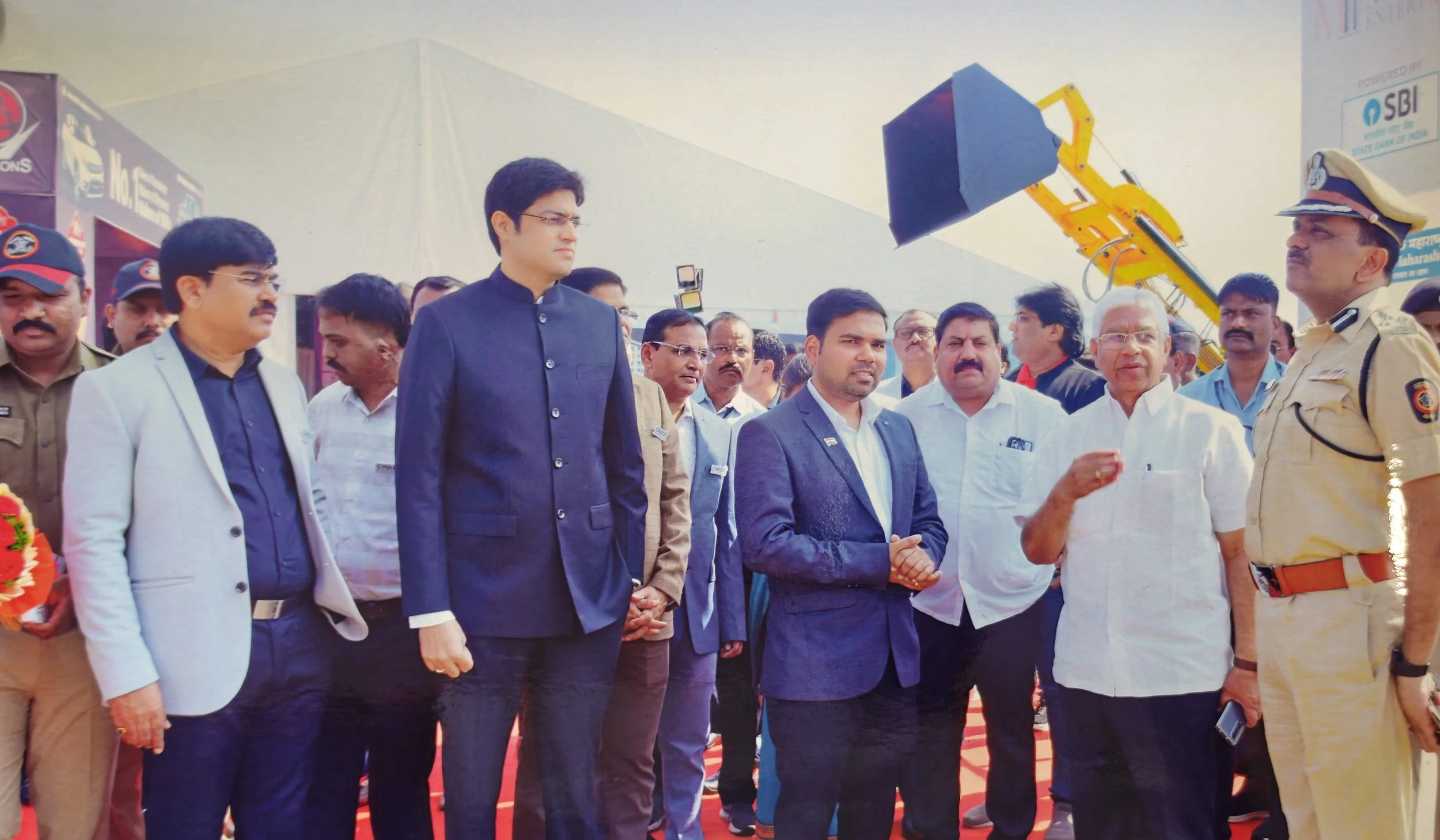 Group of officials including police officers and dignitaries attending an event with a yellow crane lifting a large black object in the background.