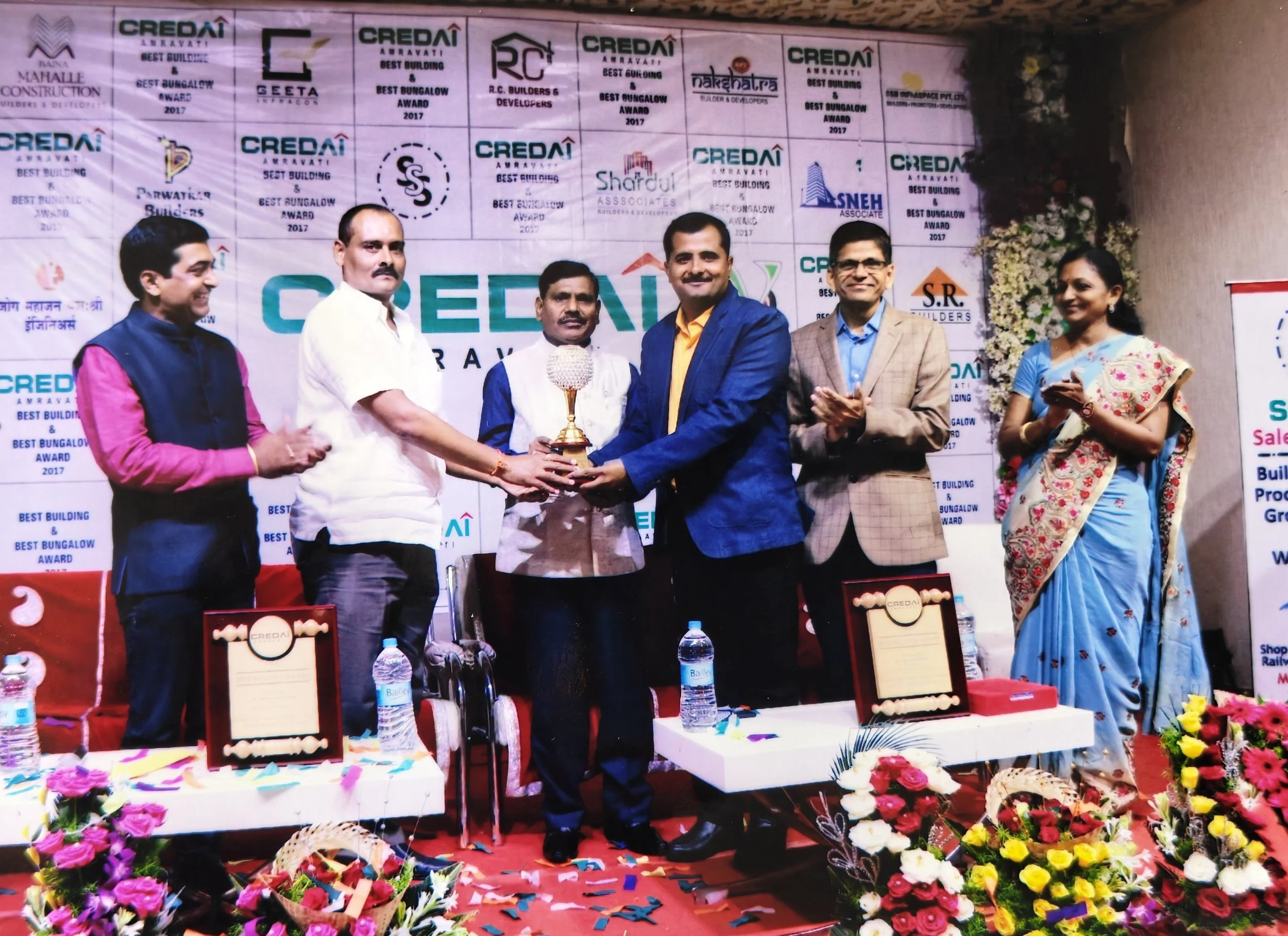 Group of six people on stage at an awards ceremony, with three men in the center exchanging a trophy, and a woman on the right clapping. The stage is decorated with flowers and banners celebrating the event.