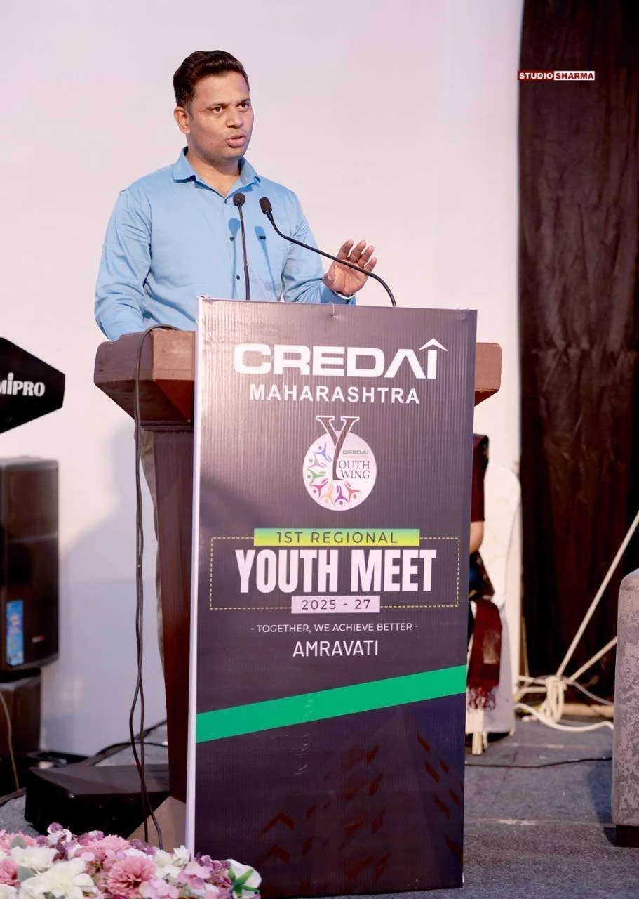 Man in blue shirt speaking at a podium during a youth meet event. The podium displays banners with logos and text for Credai Maharashtra, Youth Wing, and the event's details. The background is plain, with part of a large speaker and some decorations 