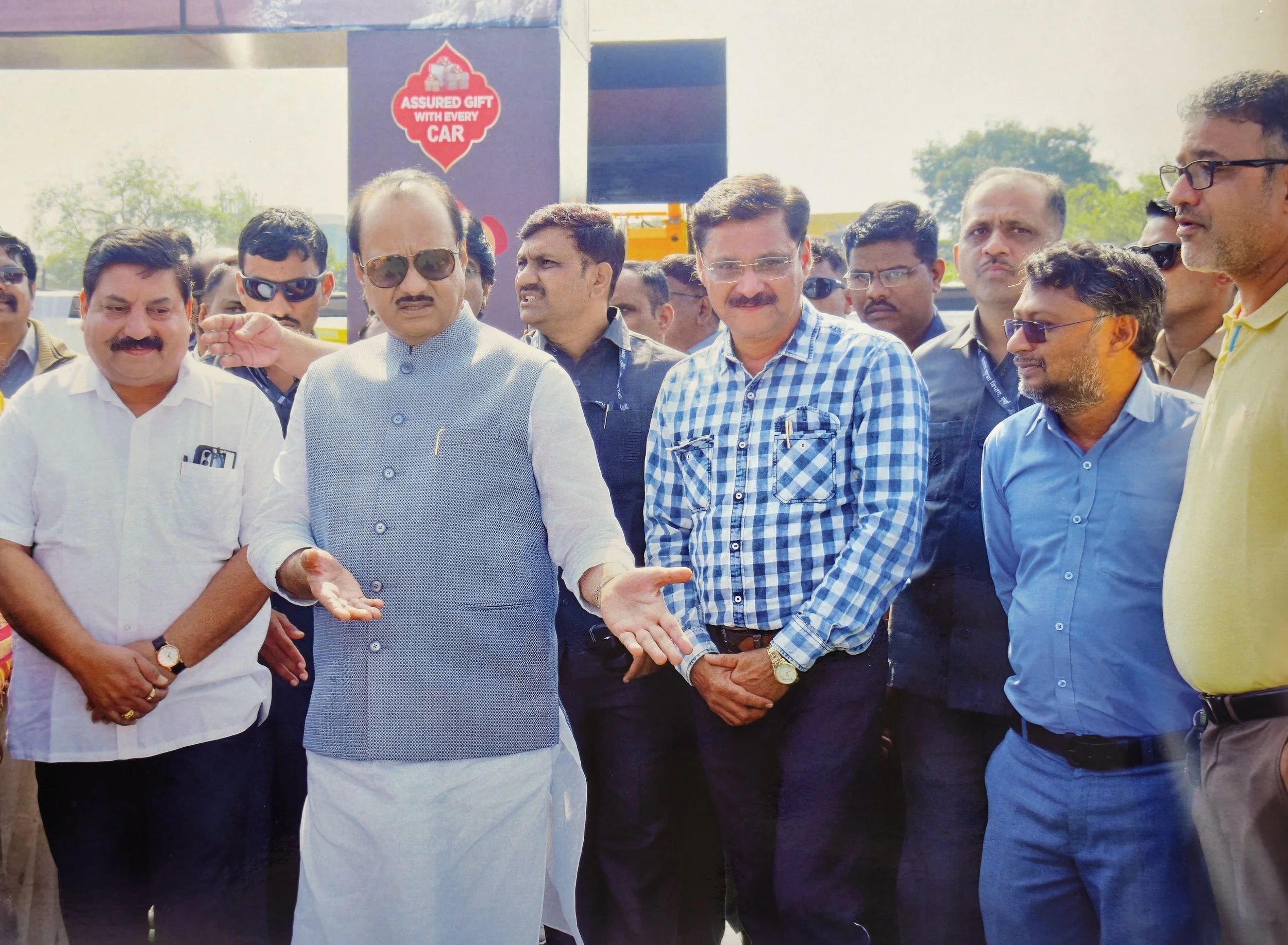 A group of men, including a man in a traditional Indian kurta and sunglasses, engaged in discussion at an outdoor event. There are several other men around, some with glasses, and behind them a sign reads 'Assured Gift with Every Car.'