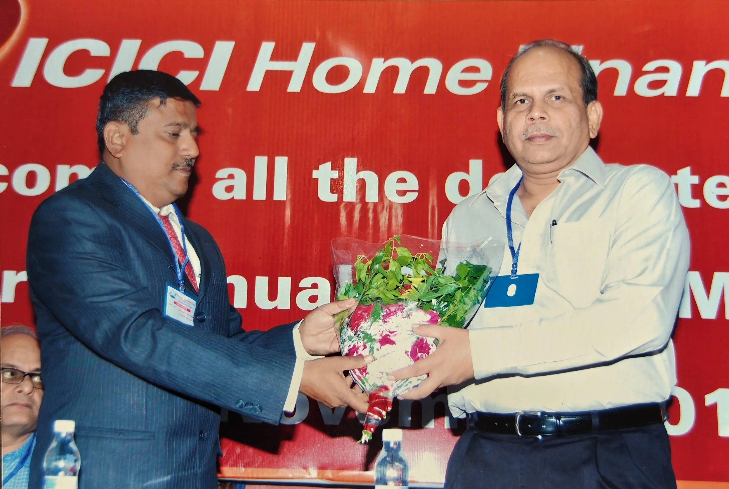 Two men at a formal event, one in a dark suit and the other in a light shirt, exchanging a bouquet of green plants wrapped in colorful paper.