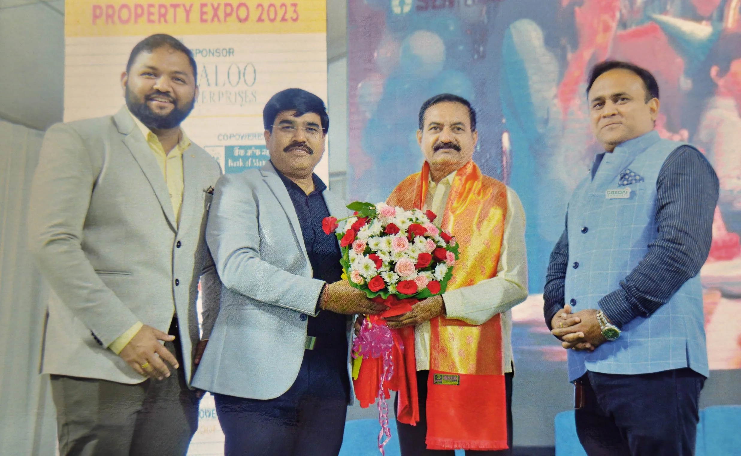 Four men in formal attire standing on stage, one holding a large bouquet of flowers, with a banner reading "Property Expo 2023" in the background.