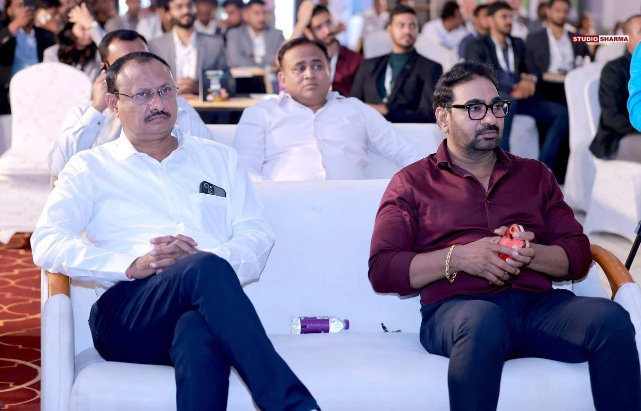 A group of men seated in a conference or seminar setting, with two men in the foreground attentively listening. The man on the left is wearing a white shirt and glasses, while the man on the right is in a maroon shirt and glasses, holding a phone. In