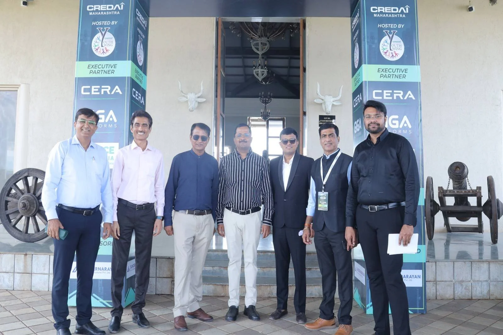 Group of seven men standing in front of a building with banners displaying 'CREDAI Maharashtra' and 'HOSTED BY Y Youth Wing.' Two mounted deer heads and a small cannon are visible on either side of the entrance.