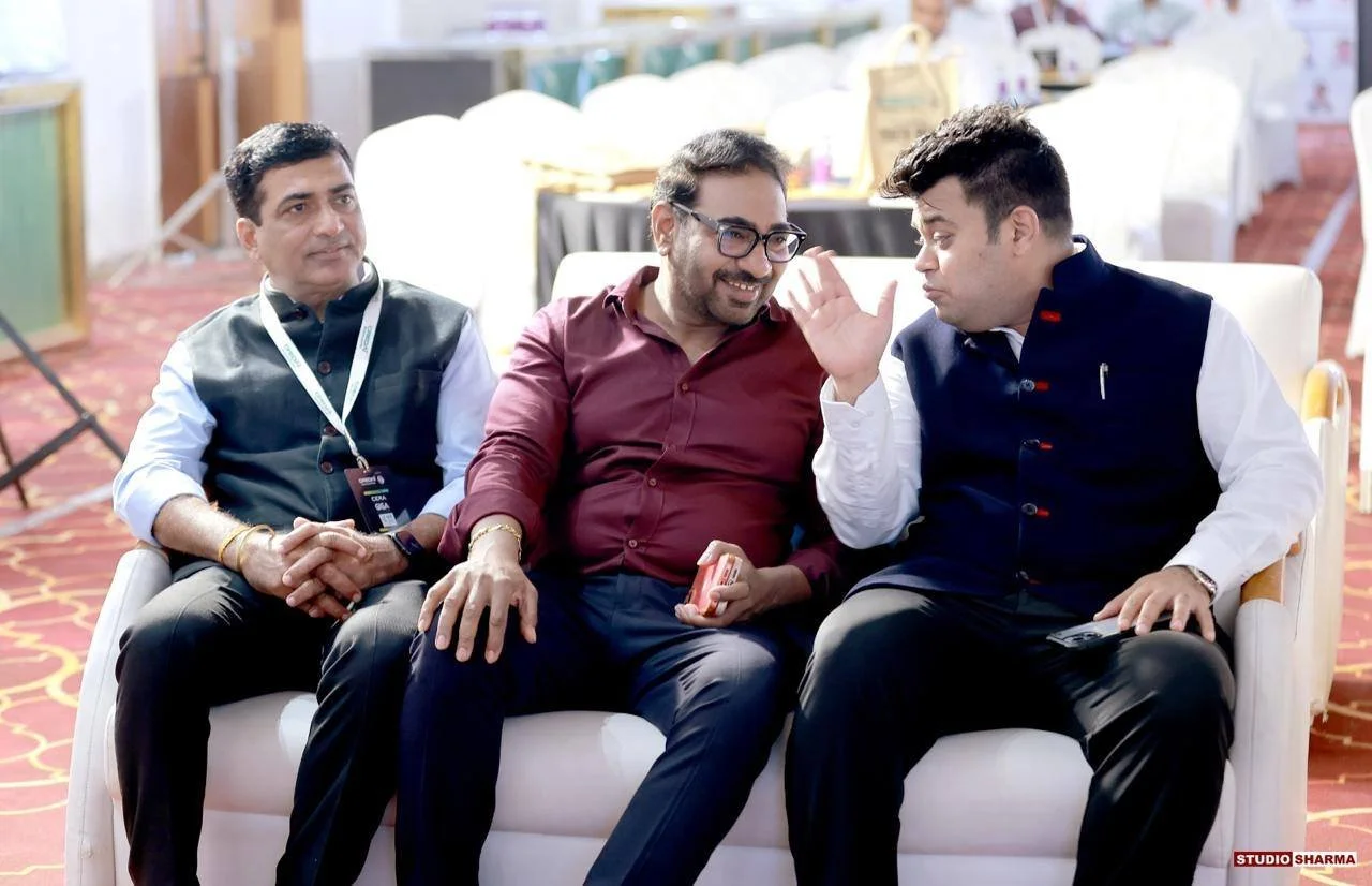 Three men sitting on a white sofa in a conference or event setting, engaged in conversation with one man whispering into another's ear. The man on the left is wearing a black vest over a white shirt, the man in the middle is wearing glasses and a mar