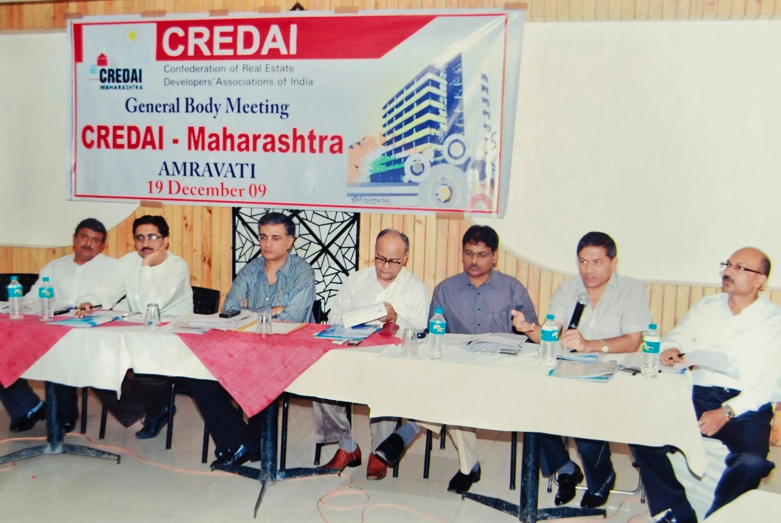 A group of seven men sitting at a long table during a formal meeting with a banner overhead that reads "CREDAI Maharashtra General Body Meeting CREDAI - Maharashtra AMRAVATI 19 December 09." The table has bottles of water, papers, and microphones.