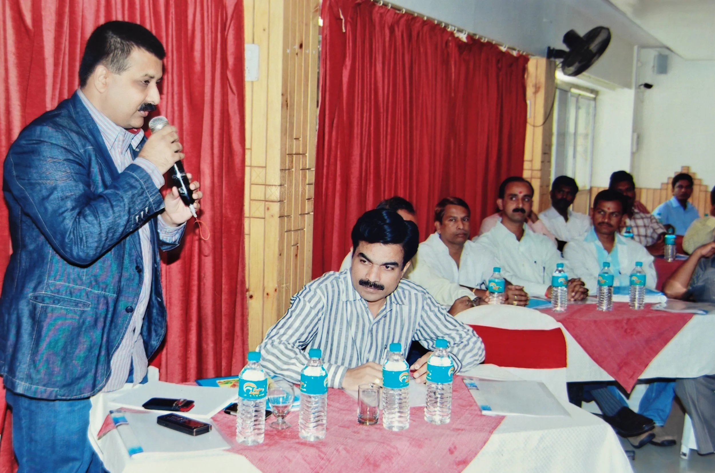 A man with a mustache and striped shirt is sitting at a table with several water bottles, a glass, and mobile phones, while another man in a blue jacket and striped shirt is speaking into a microphone during a meeting or conference.