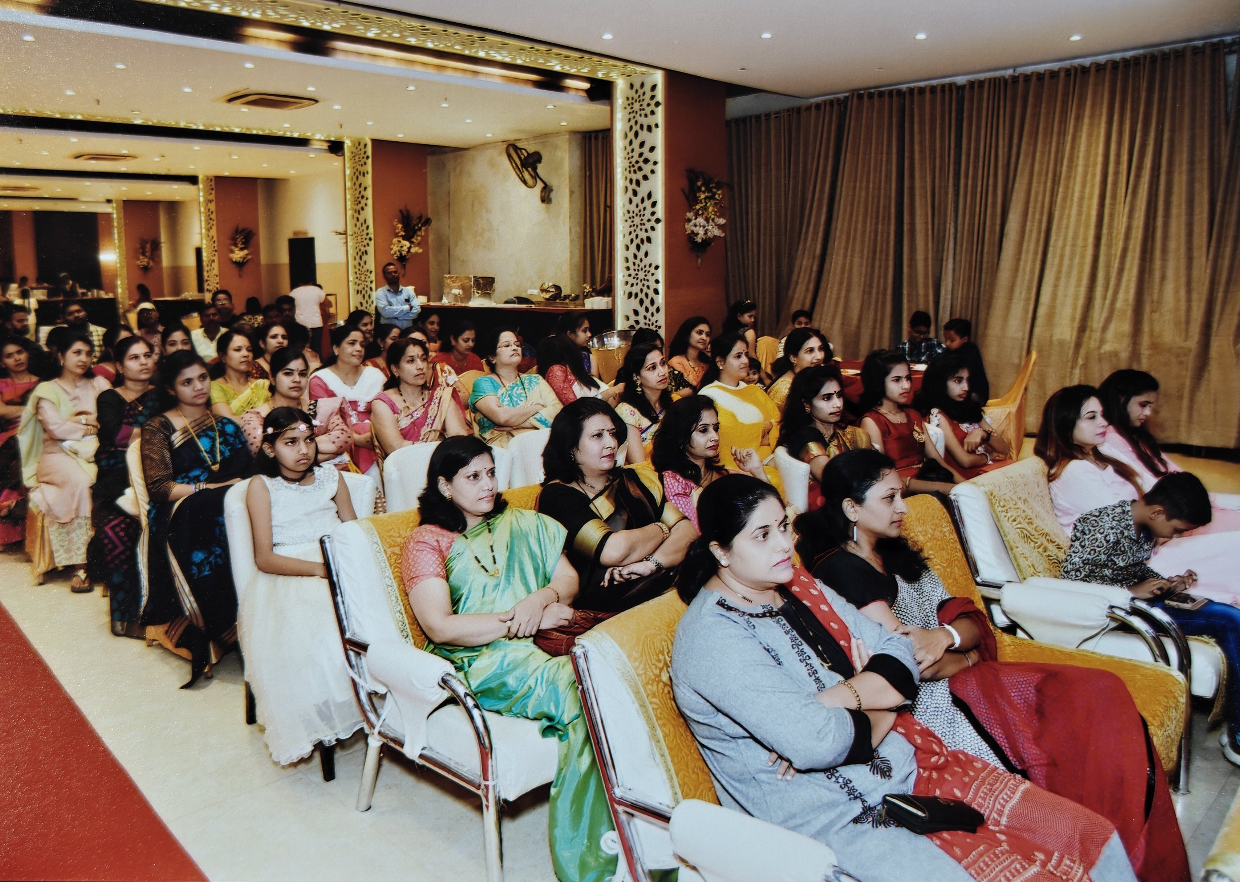 Women and children attending an indoor event, seated on chairs, paying attention to a performance or presentation. The women are dressed in colorful traditional Indian attire.
