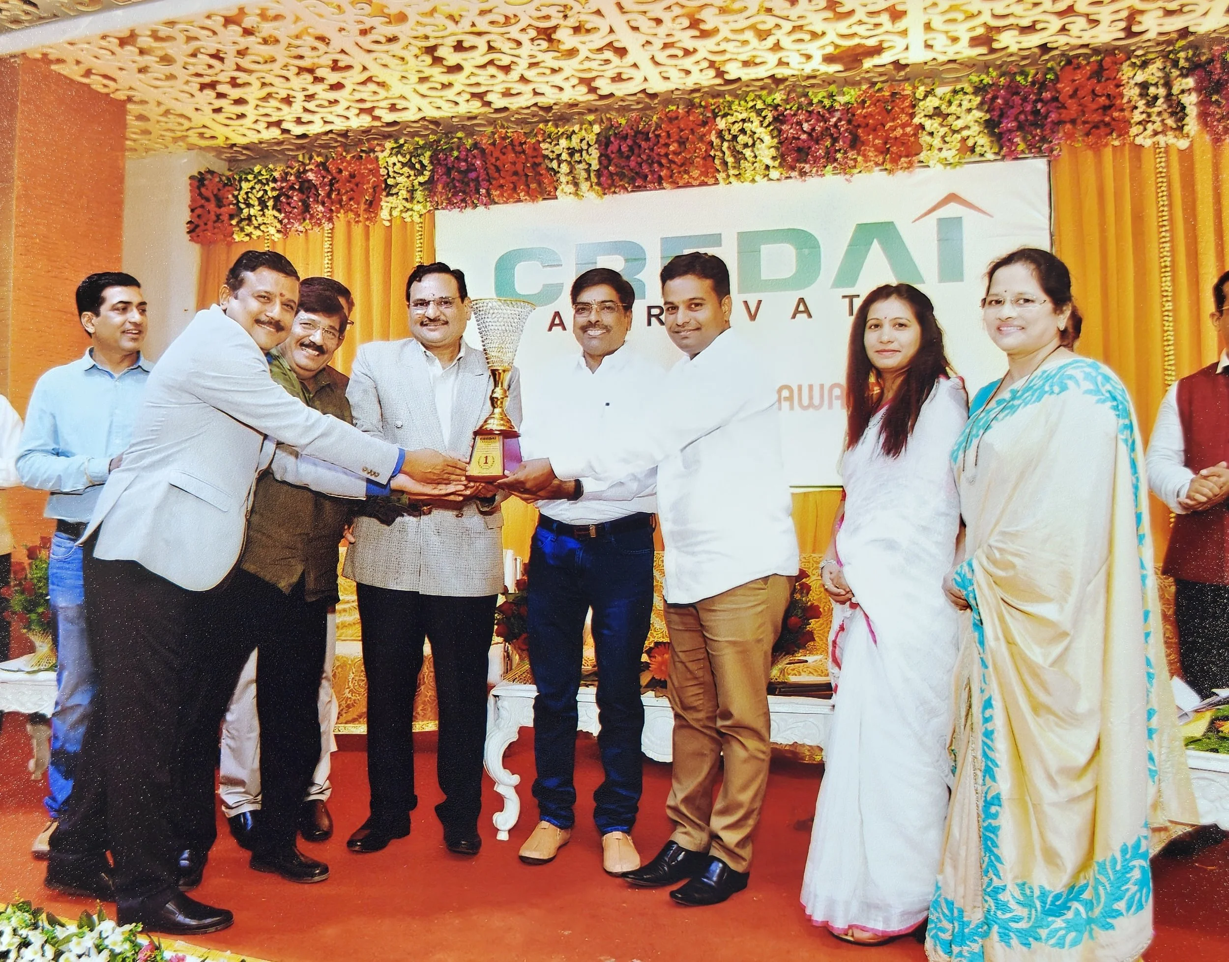 Group of people at an award ceremony, with some presenting and receiving a trophy on stage decorated with colorful flowers and a banner in the background that says "GRAI" and "AWARD".
